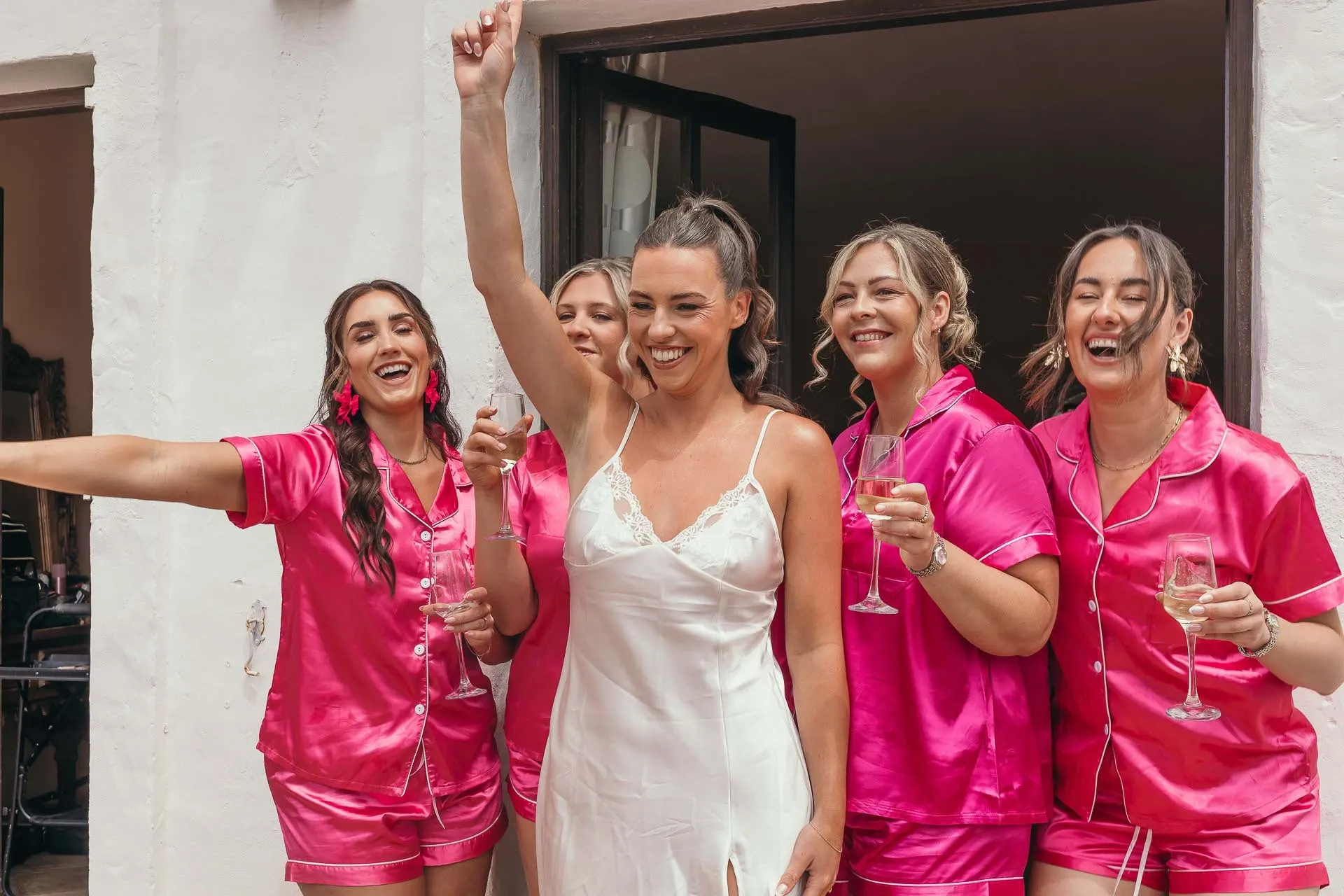 Bride in a white slip celebrating with bridesmaids in pink pyjamas, glasses raised, Hacienda San José, Mijas