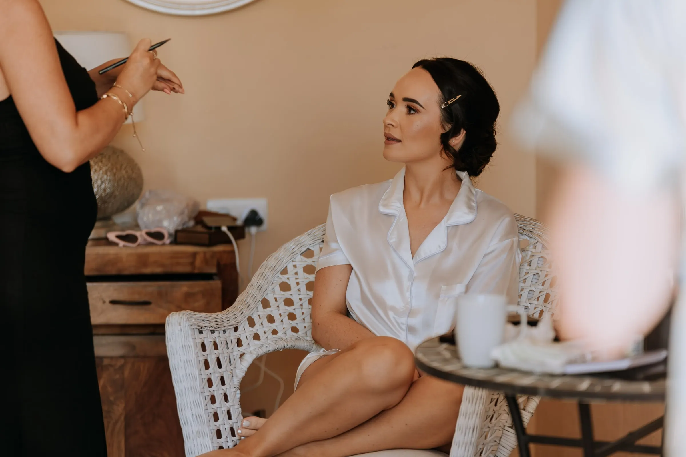 Bride in white pyjamas having her make-up done in a wicker chair, wedding morning in Mijas