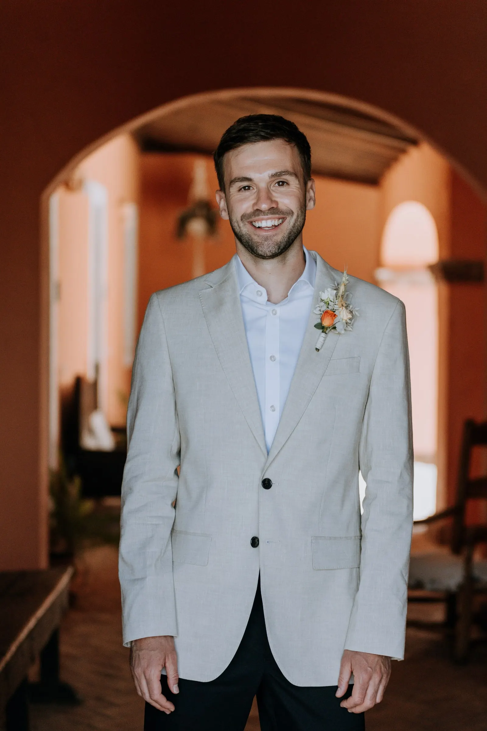 Portrait of the smiling groom under an arch in a beige jacket and peach rose buttonhole, Mijas