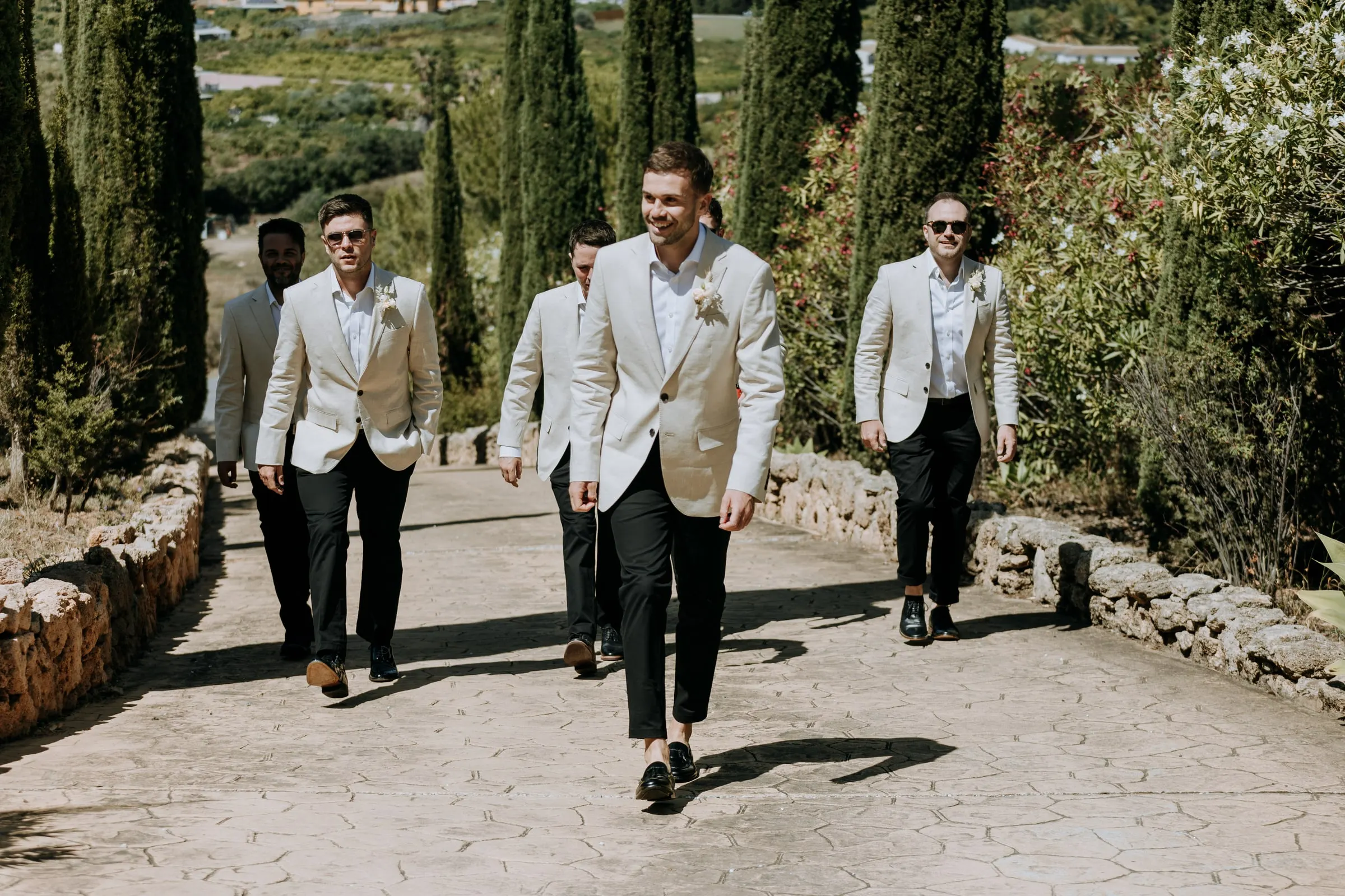 Groom and groomsmen in beige jackets walking up the cypress avenue of Hacienda San José, Mijas