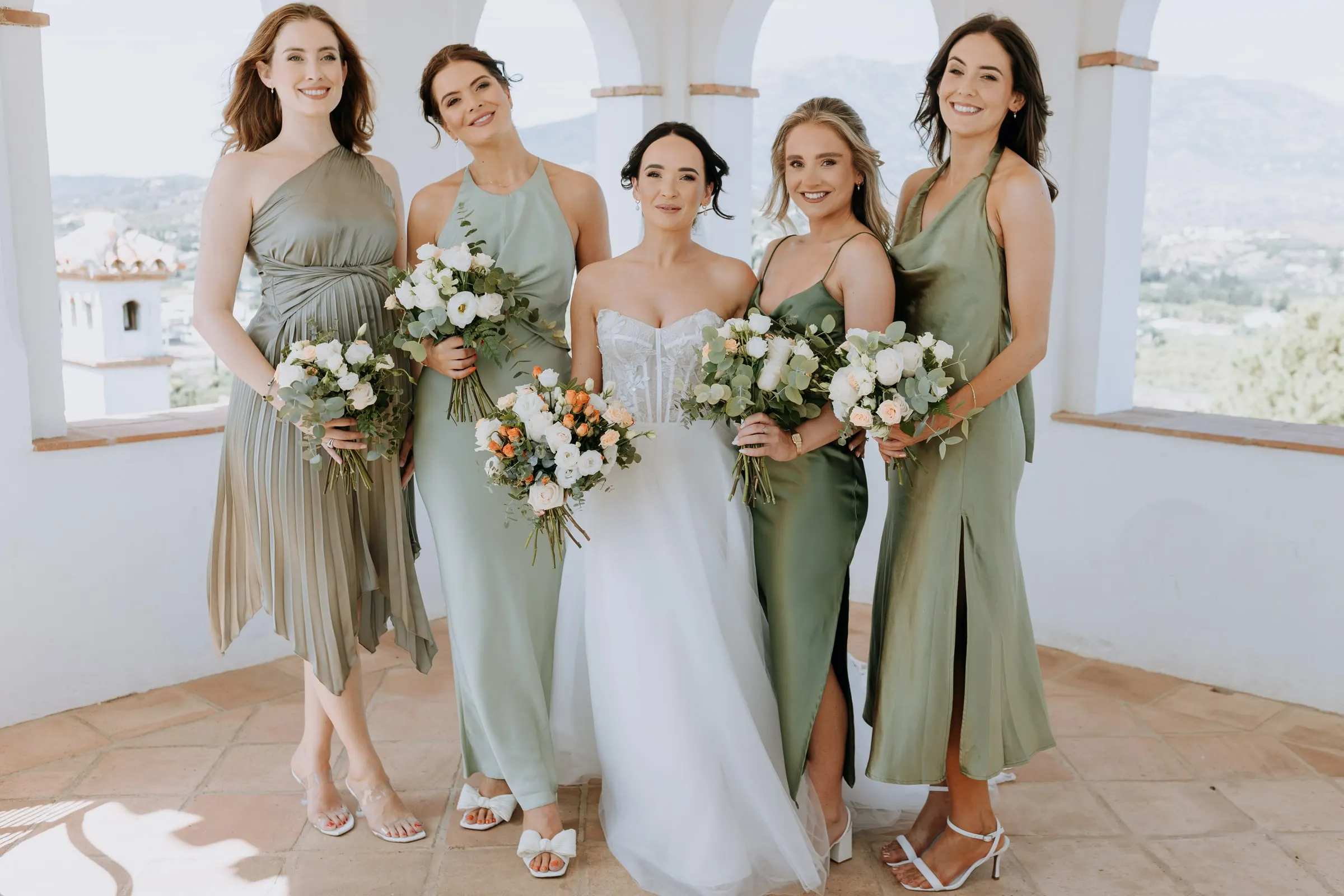 Bride with her bridesmaids in different shades of sage green under the tower arches, Hacienda San José