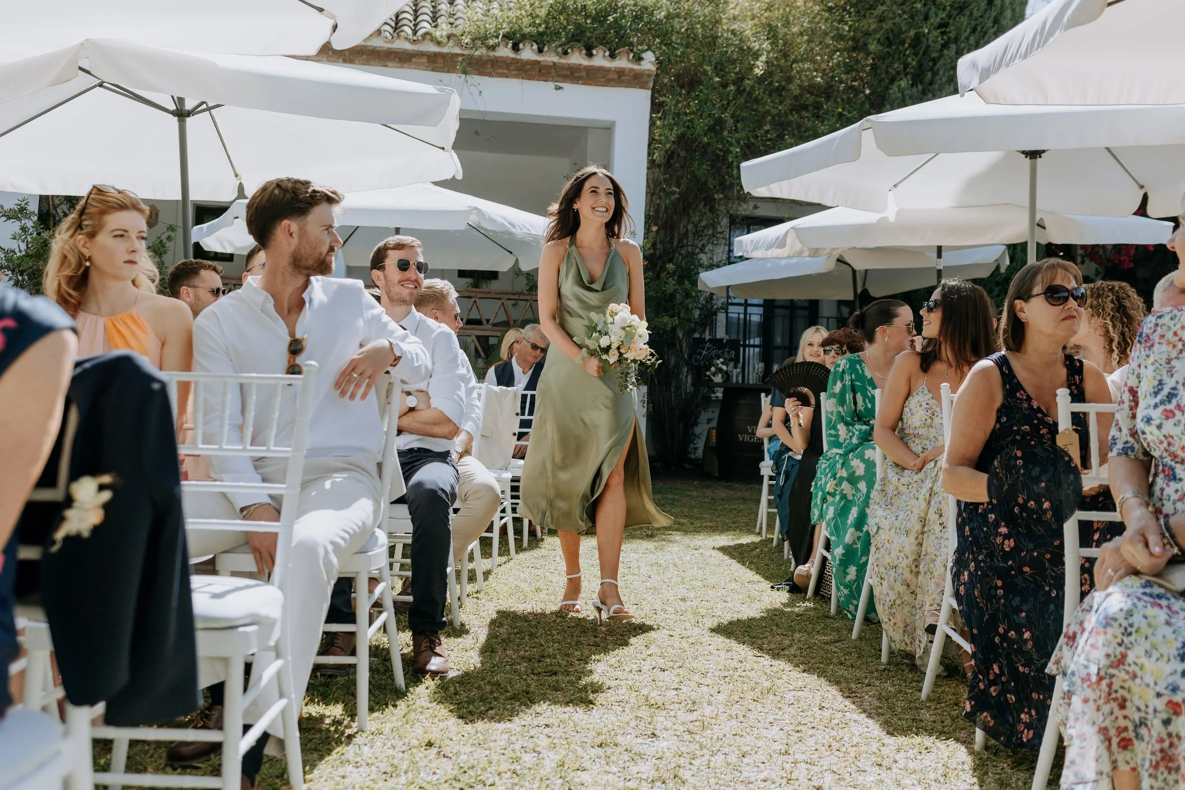 Bridesmaid in sage green walking down the lawn aisle as guests turn, Hacienda San José