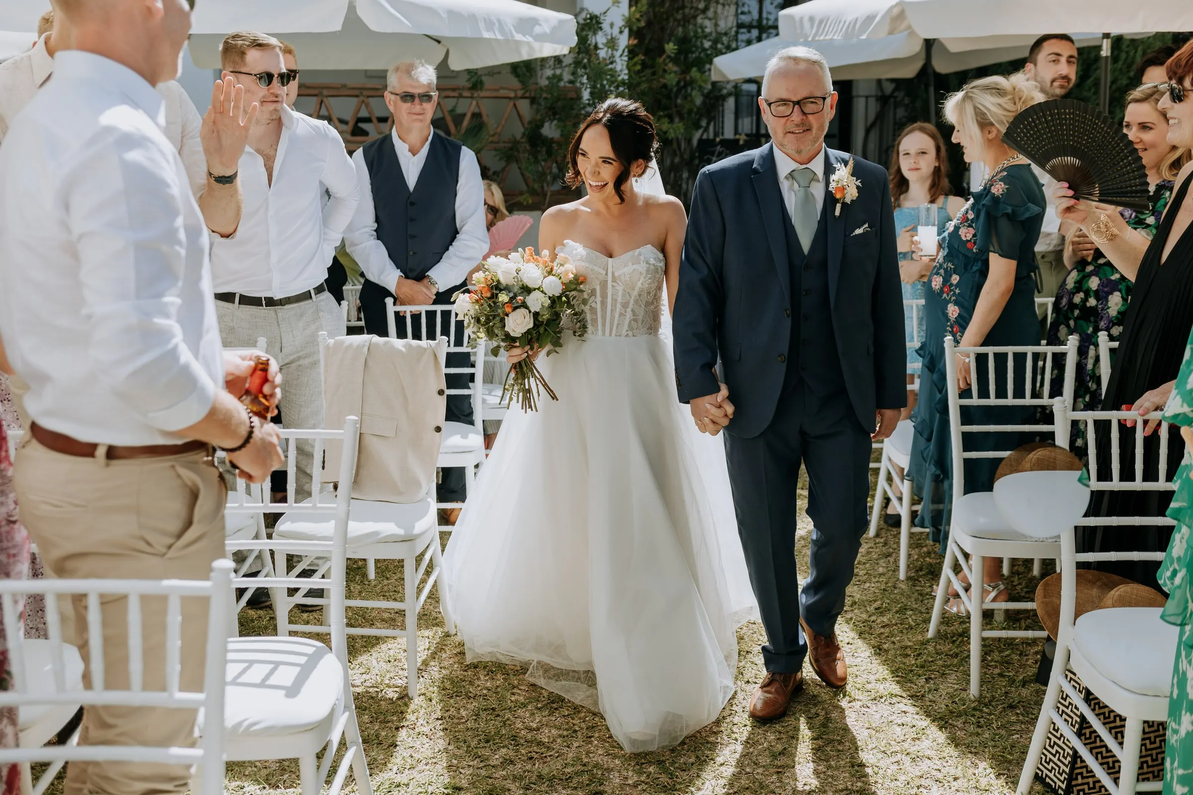 Bride and father between the white chairs as guests watch, garden ceremony in Mijas