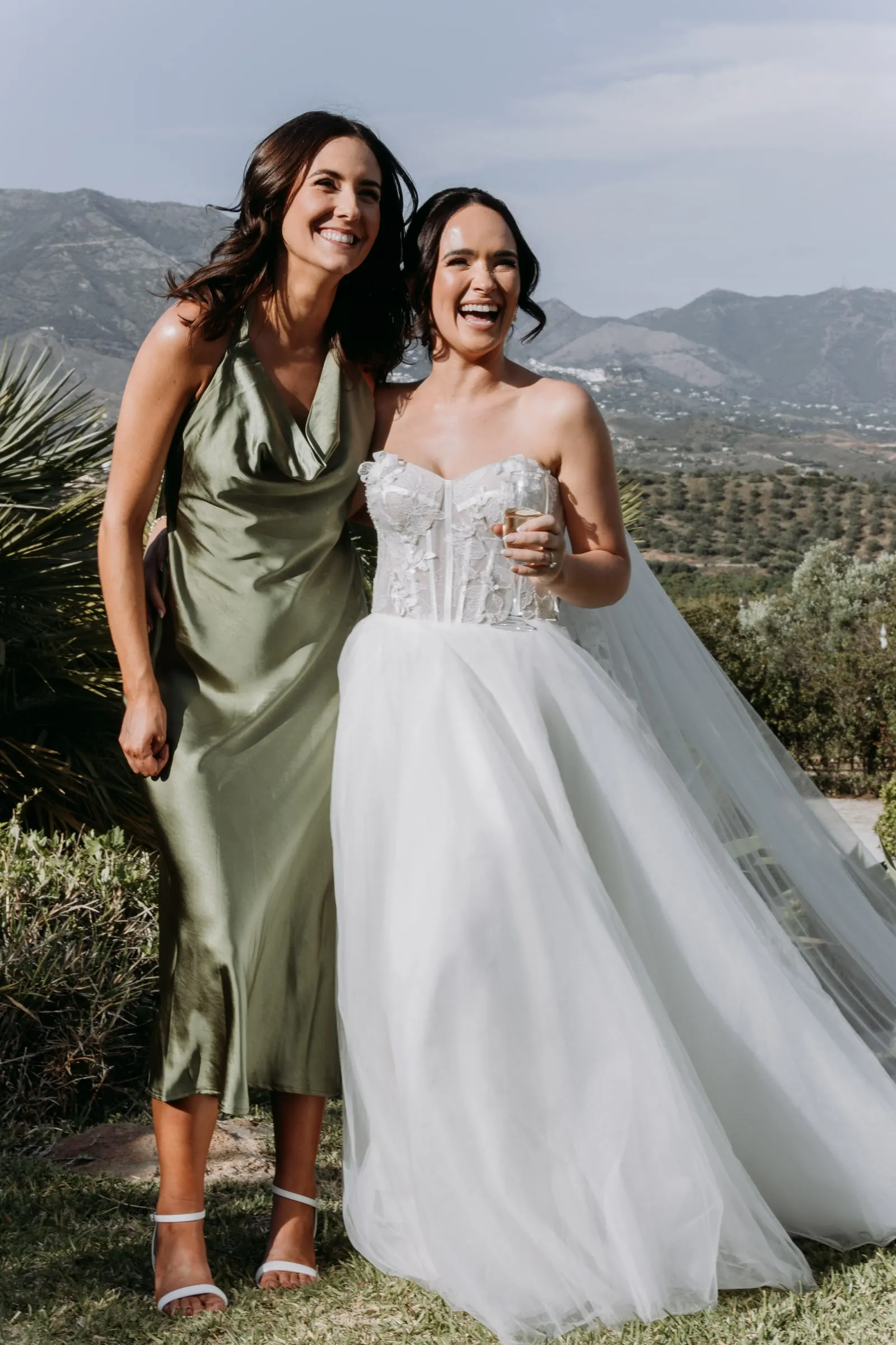 Bride laughing with a friend in olive satin, with the hills of Mijas behind