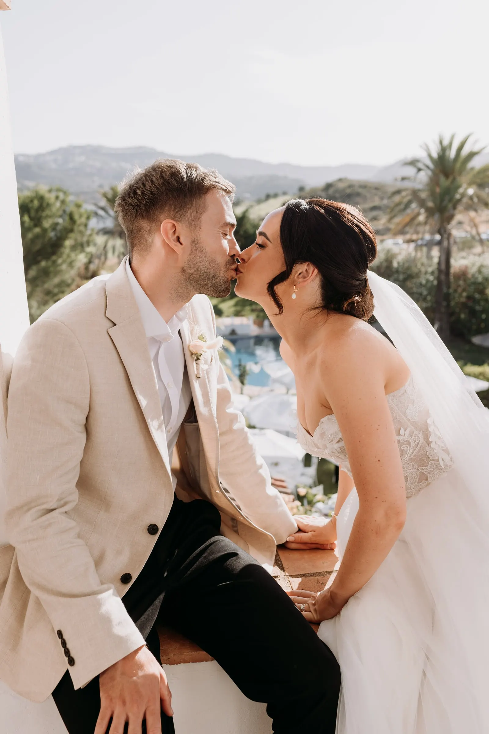Bride kissing the groom on the cheek on the tower ledge, with palms and mountains behind, Mijas