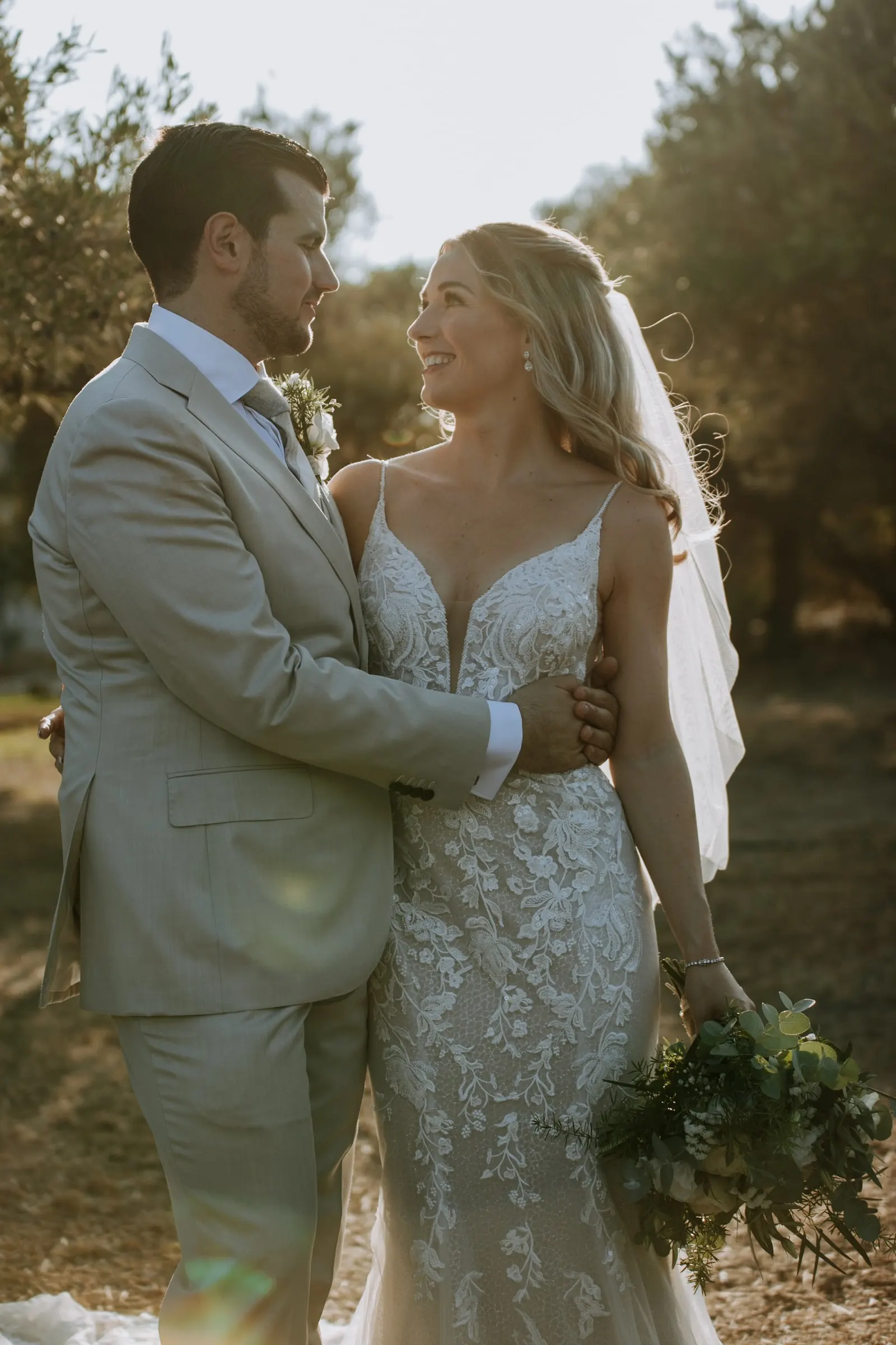 Bride and groom embracing in golden evening light among olive trees, bouquet in hand, Hacienda San José, Mijas