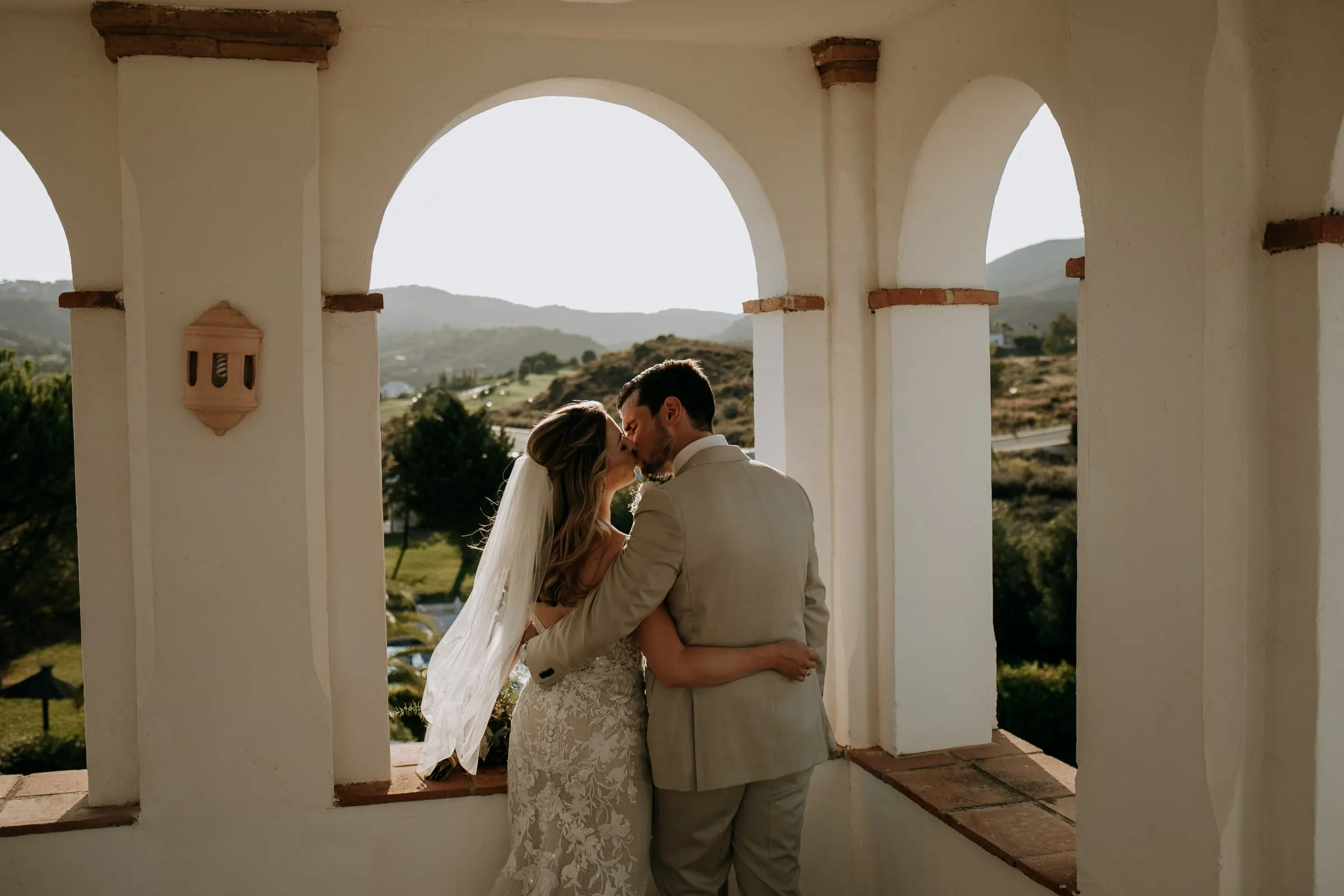 Bride and groom kissing under a white arch of the loggia, hills beyond, Hacienda San José, Mijas