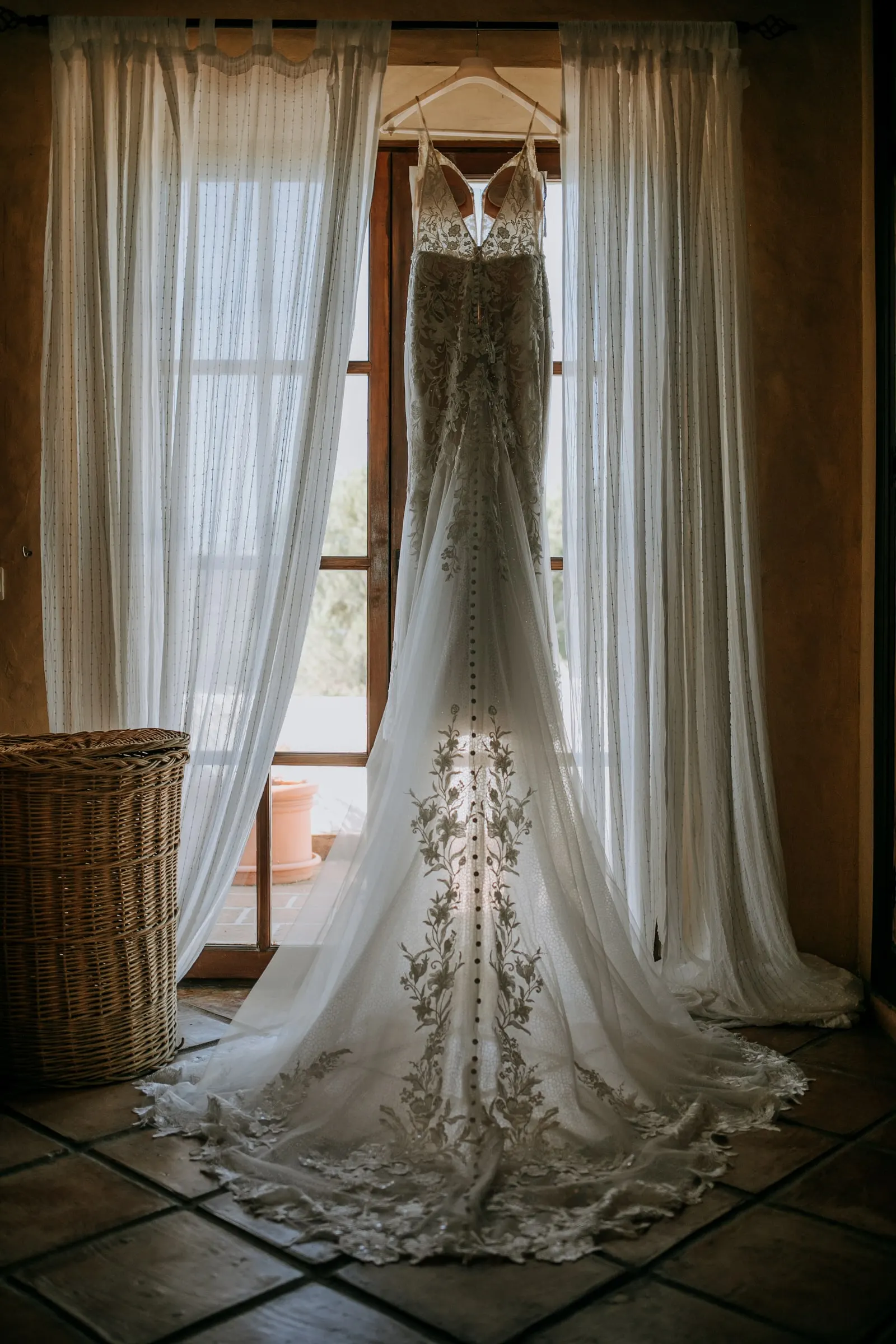 Lace wedding dress with a long train hanging in front of a curtained window, Hacienda San José, Mijas