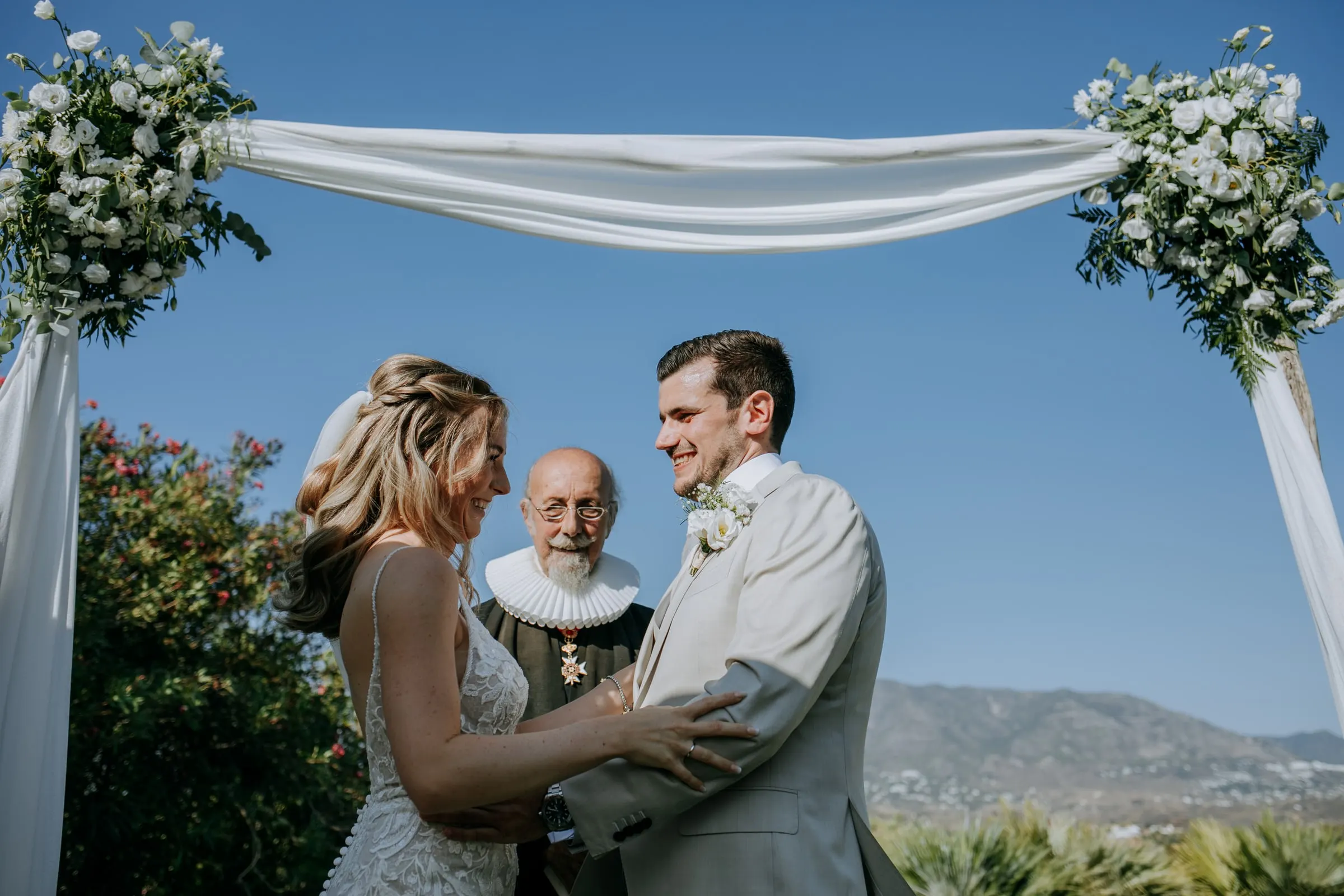Bride and groom hold hands before the officiant under a flower-draped arch, mountains behind, Hacienda San José, Mijas