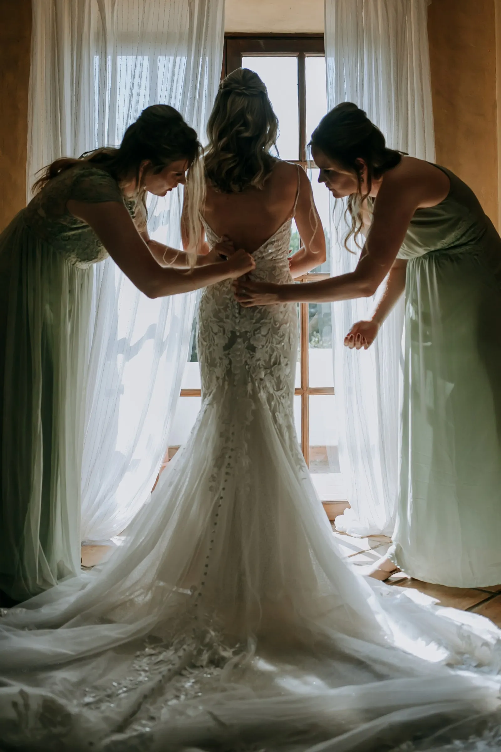 Two bridesmaids in green lace up the back of the bride's dress against a bright window, Hacienda San José, Mijas