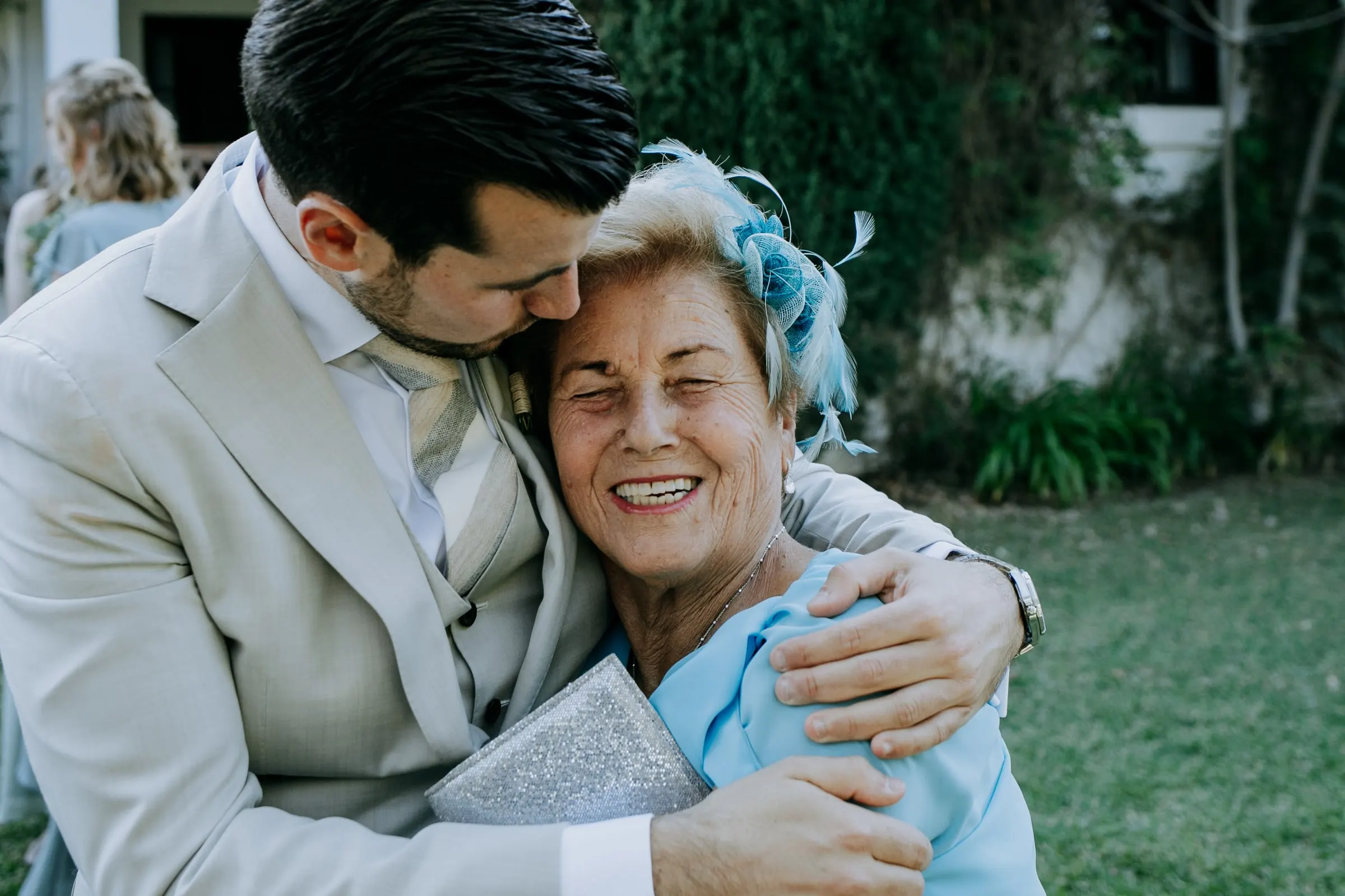 Groom hugging a smiling older guest in a blue fascinator on the lawn, Hacienda San José, Mijas