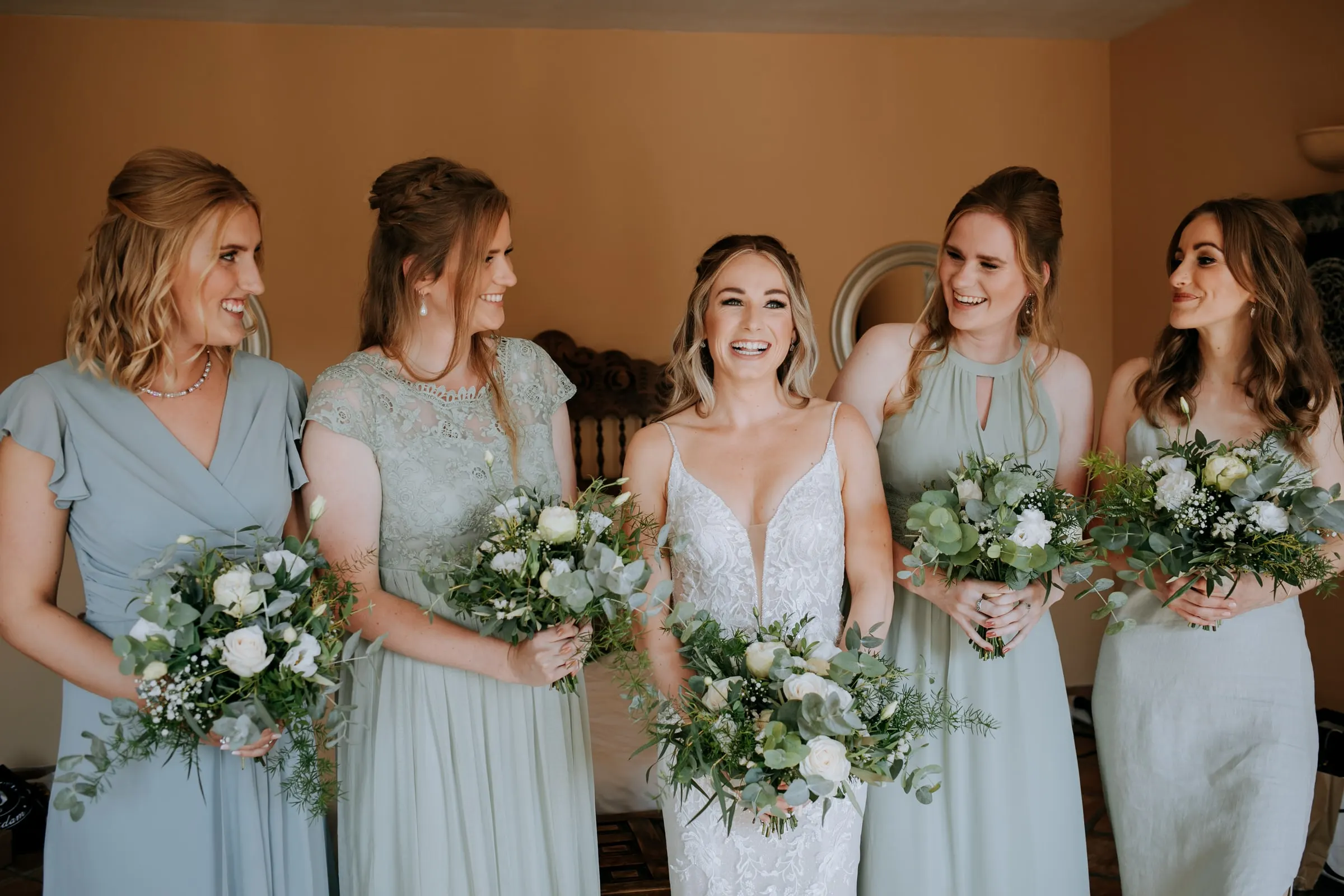Bride laughing with four bridesmaids in sage and blue dresses, all holding white bouquets, Hacienda San José, Mijas