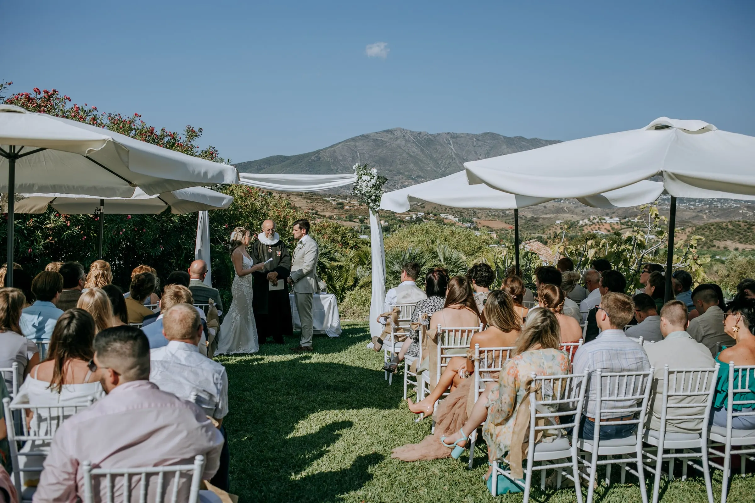 Guests seated on the lawn under white parasols facing the ceremony, mountains behind, Hacienda San José, Mijas