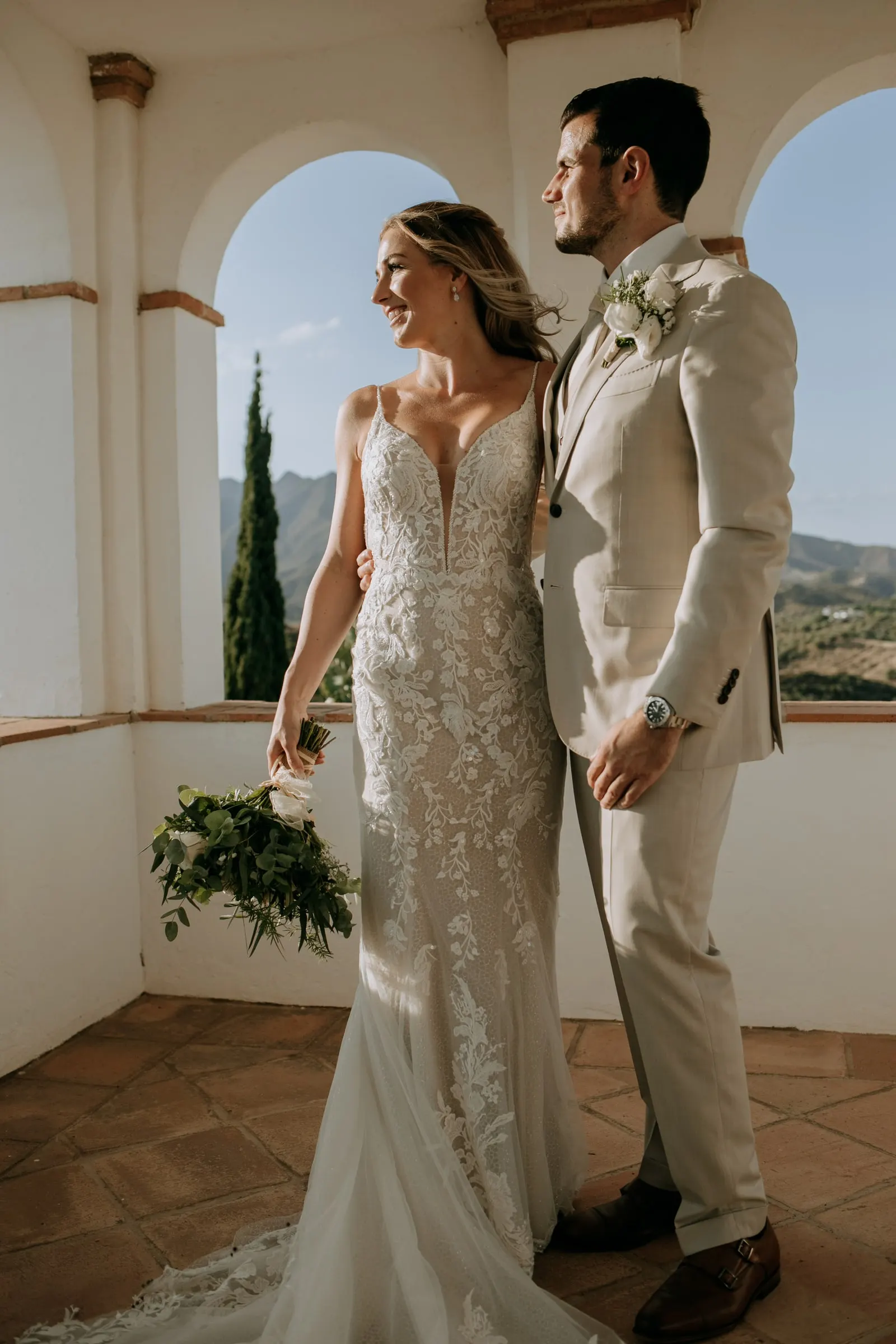 Bride and groom smiling in the arched loggia, her bouquet held low, hills behind, Hacienda San José, Mijas