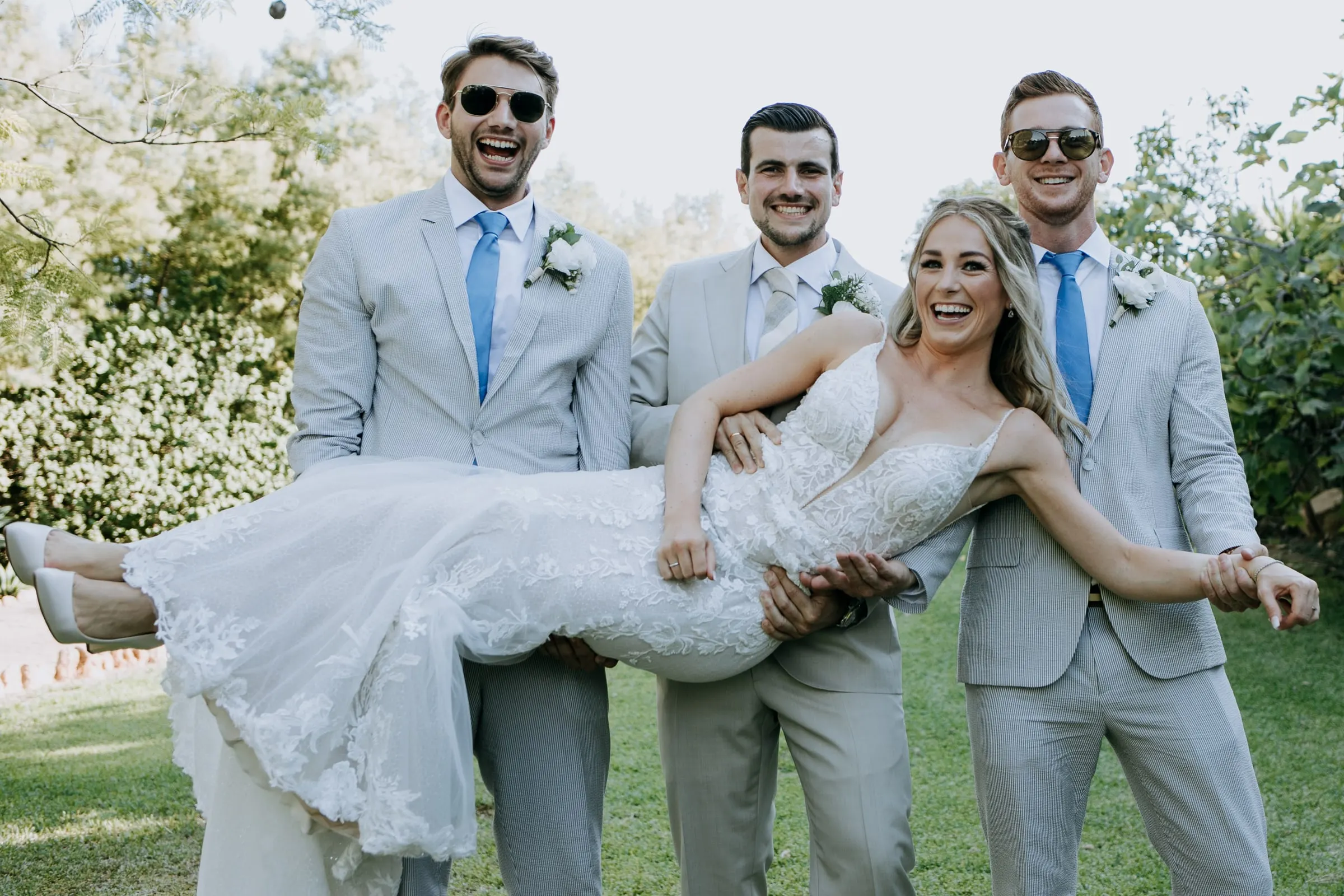 Three groomsmen in grey suits and sunglasses lift the laughing bride on the lawn, Hacienda San José, Mijas