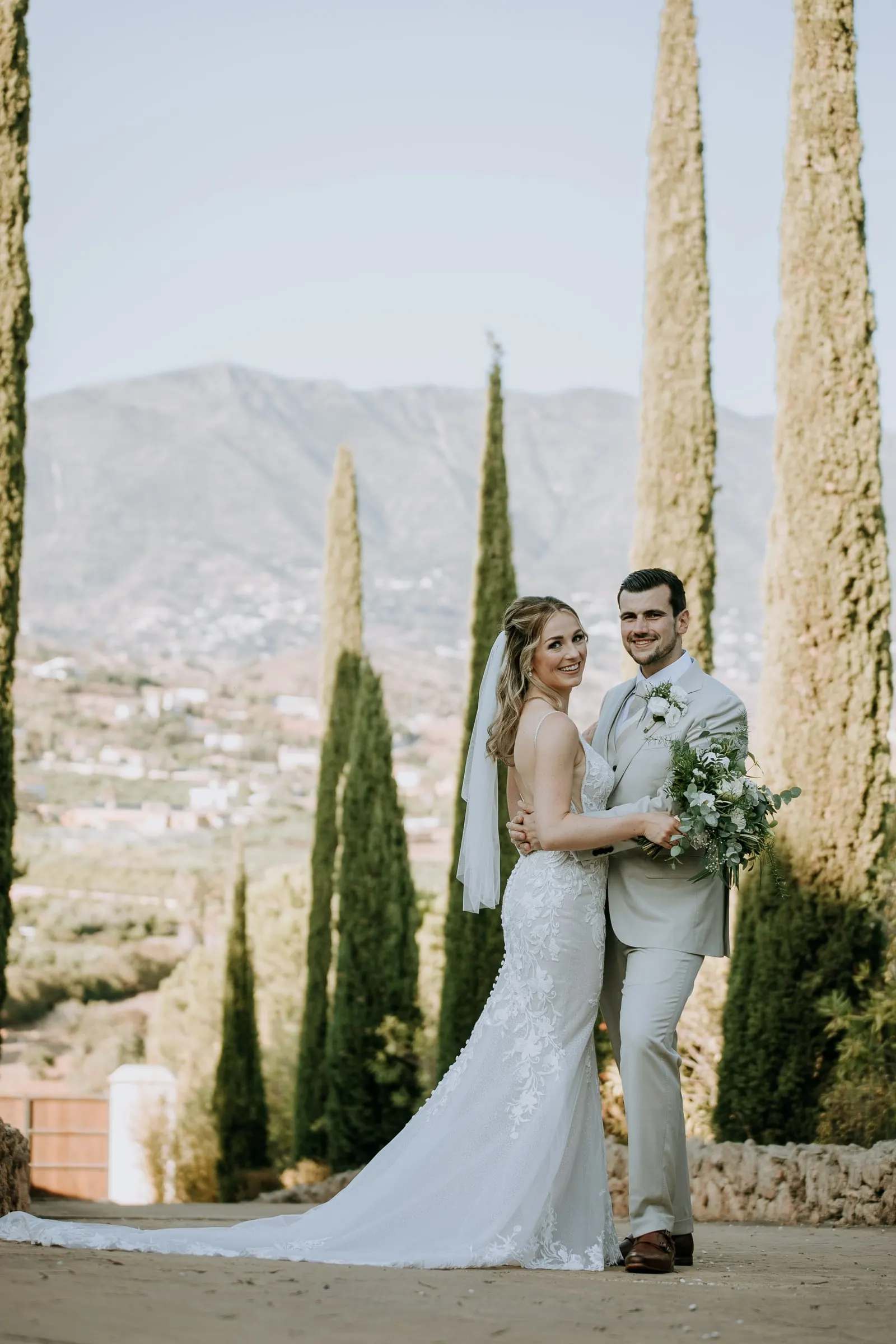 Bride and groom posing between tall cypresses with the mountains behind, her train on the path, Hacienda San José, Mijas