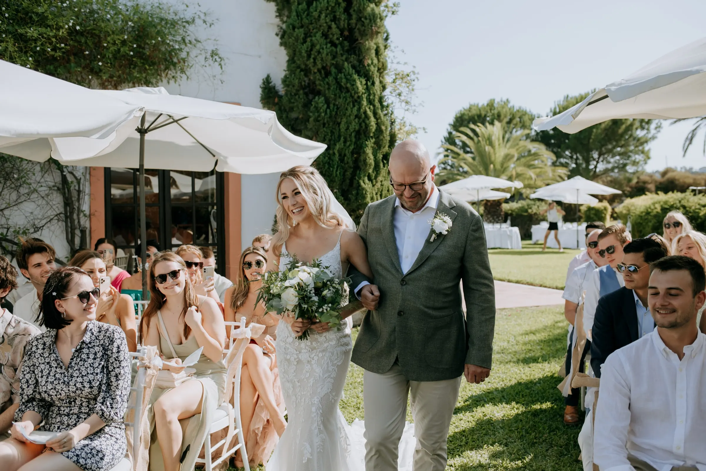 Bride walking down the aisle on a man's arm as guests in sunglasses turn to watch, Hacienda San José, Mijas