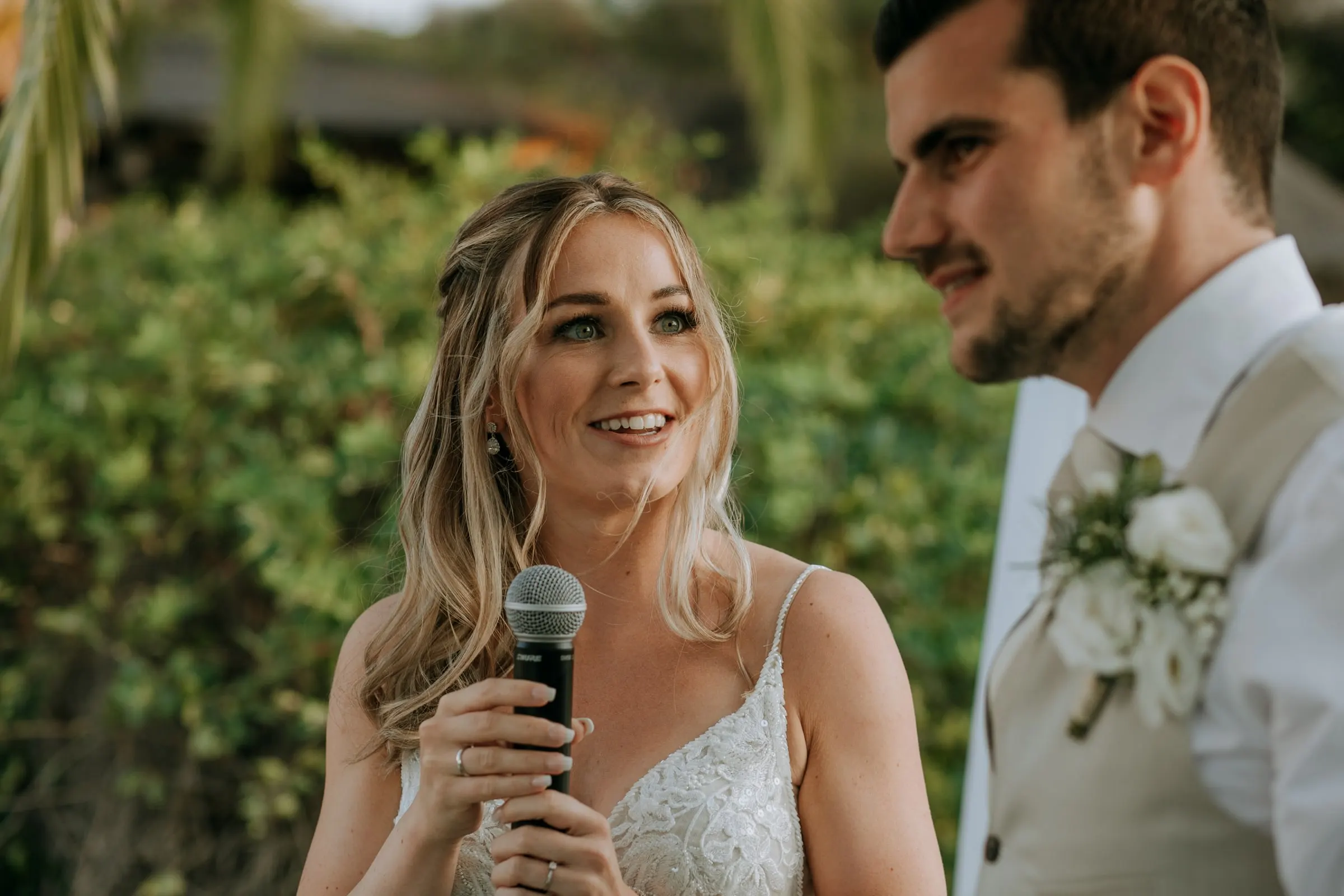 Bride giving a speech into a microphone as the groom smiles at her side, Hacienda San José, Mijas