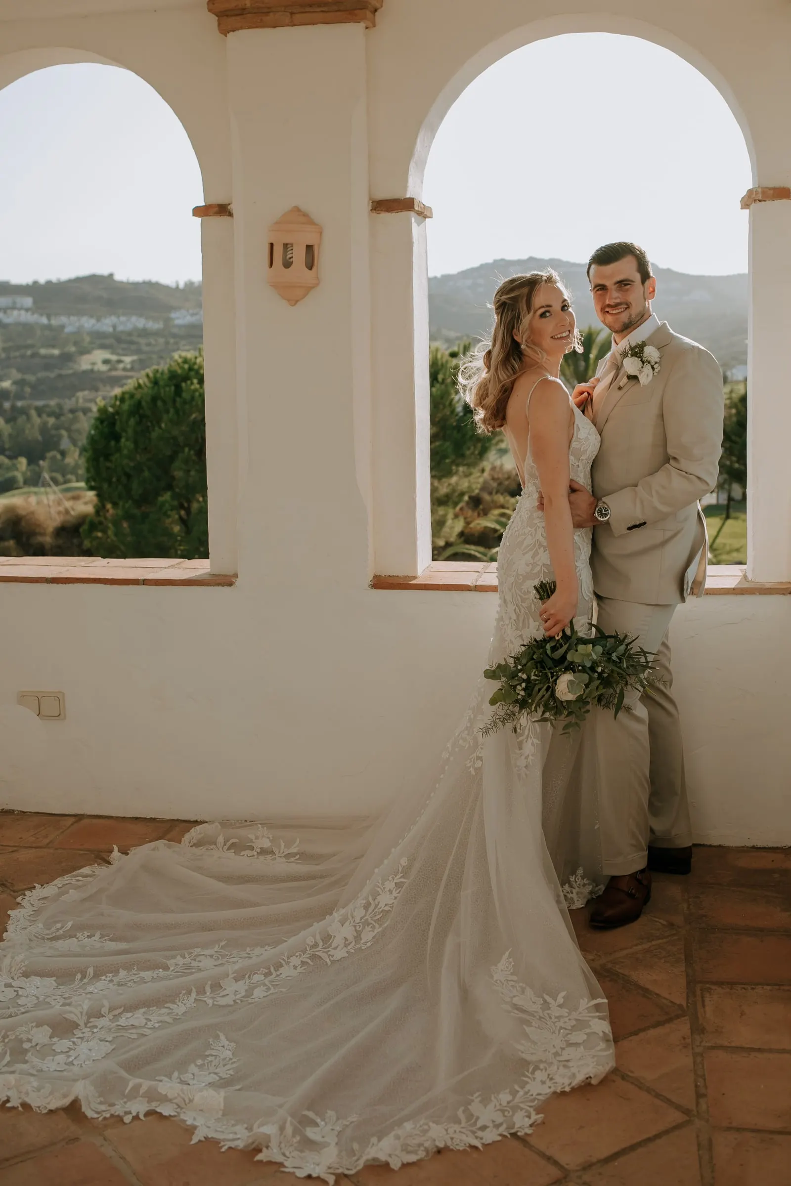 Bride and groom framed by a white arch, her lace train spread over the terracotta floor, Hacienda San José, Mijas
