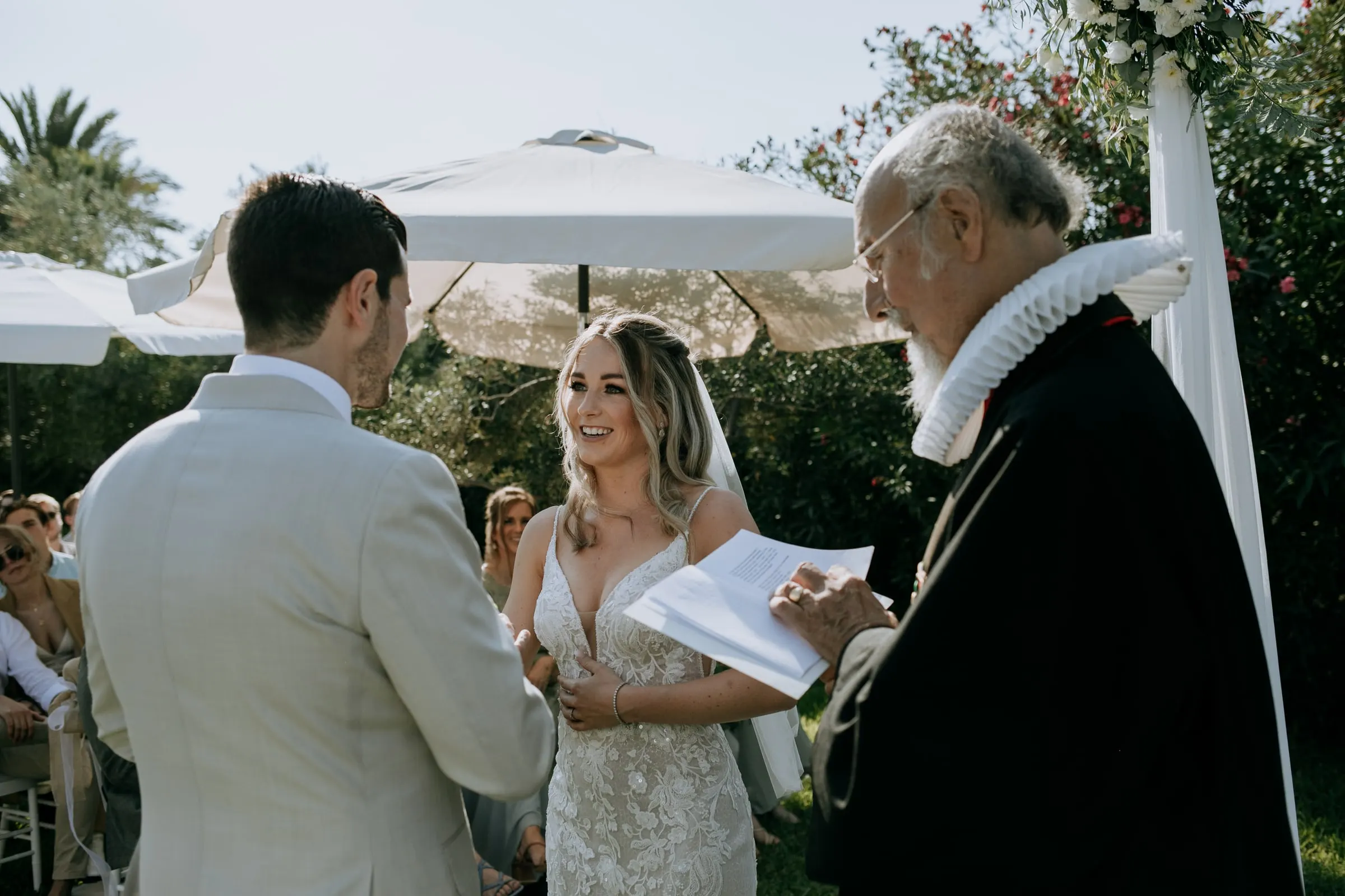 Bride smiling as the officiant in a ruffled collar reads during the ceremony, Hacienda San José, Mijas