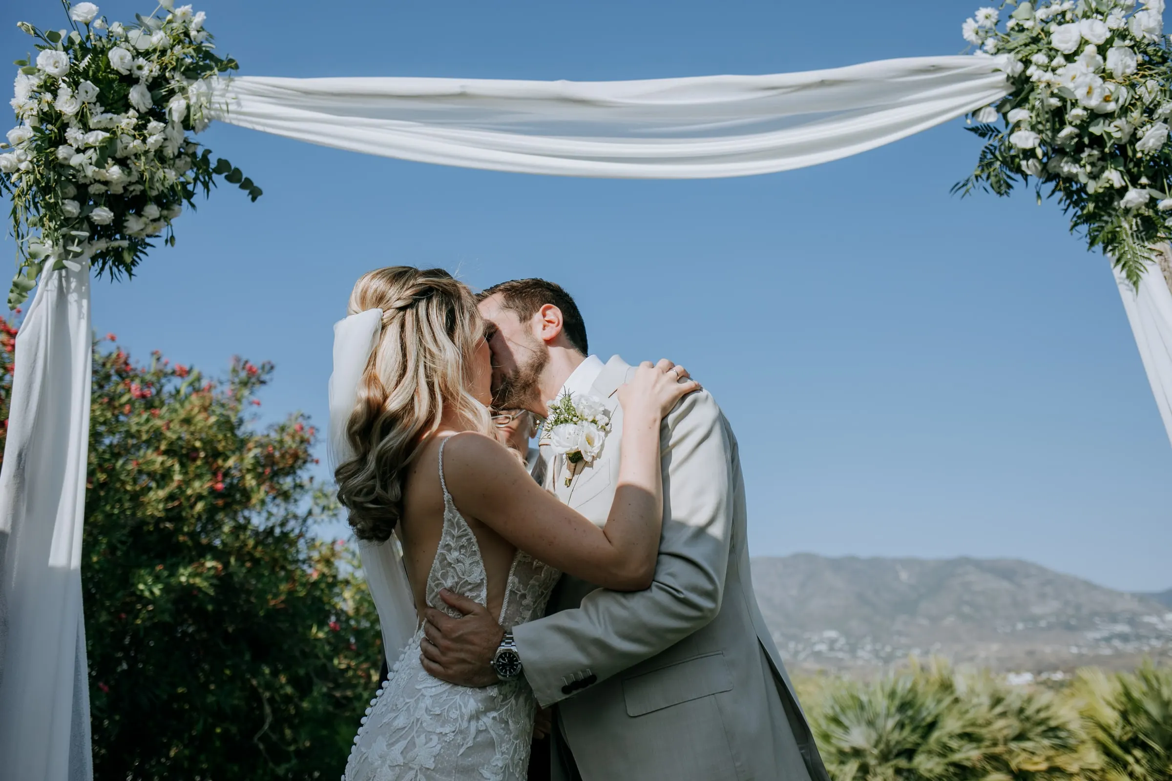 Bride and groom kiss under the flower-draped ceremony arch against a blue sky, Hacienda San José, Mijas