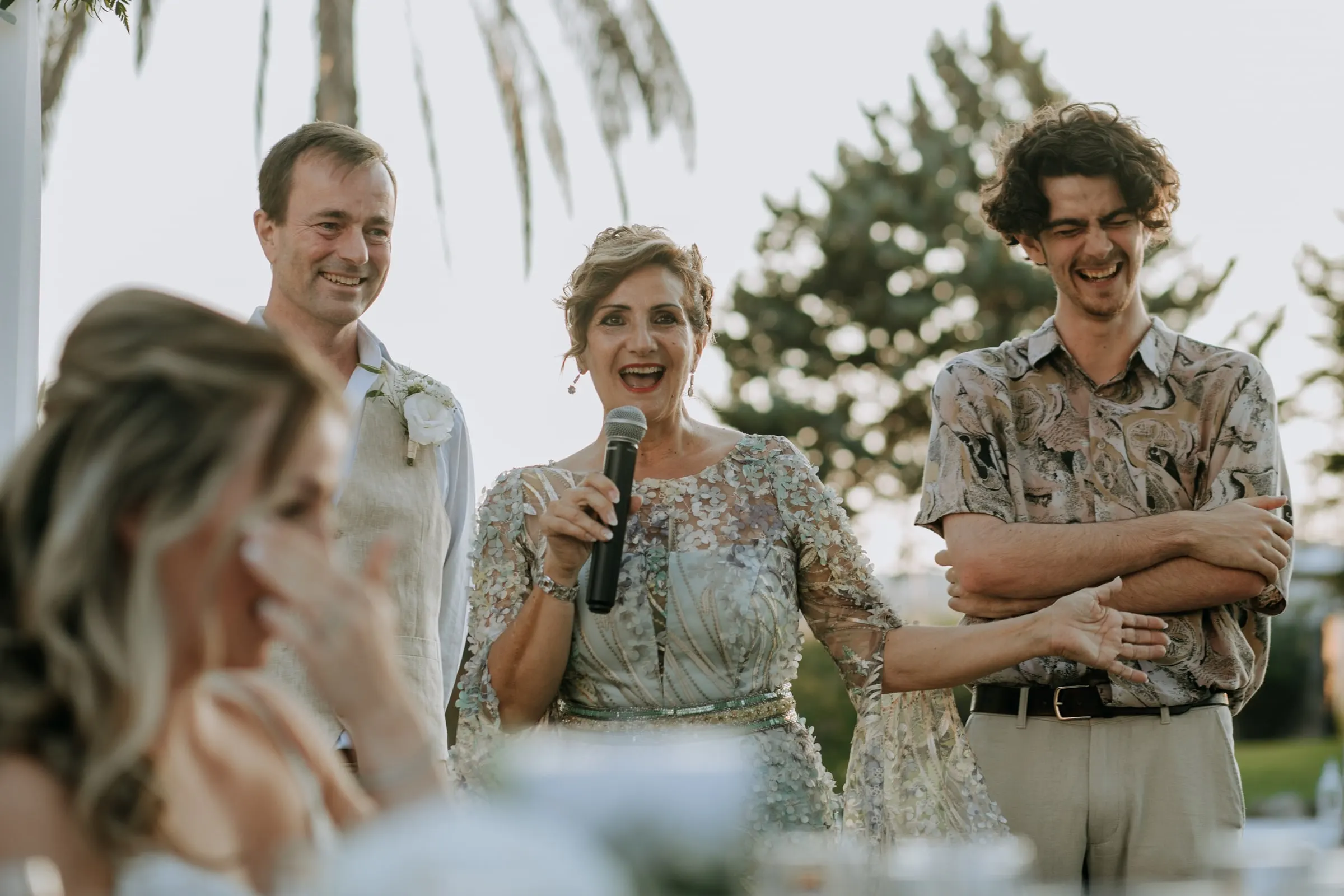 A woman giving a laughing speech into a microphone as the bride wipes away a tear, Hacienda San José, Mijas