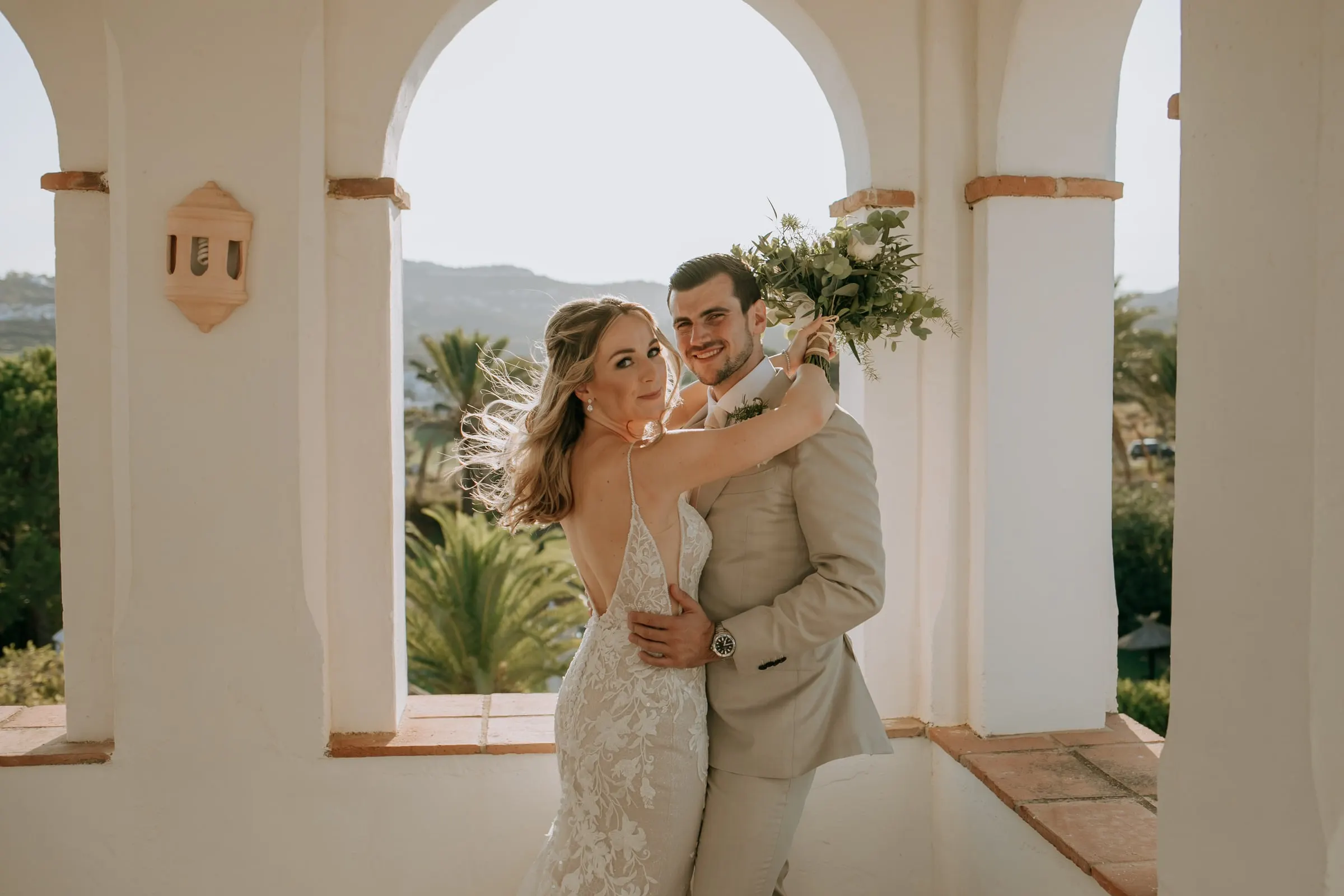 Bride and groom embracing under the white arches, bouquet raised in the breeze, Hacienda San José, Mijas