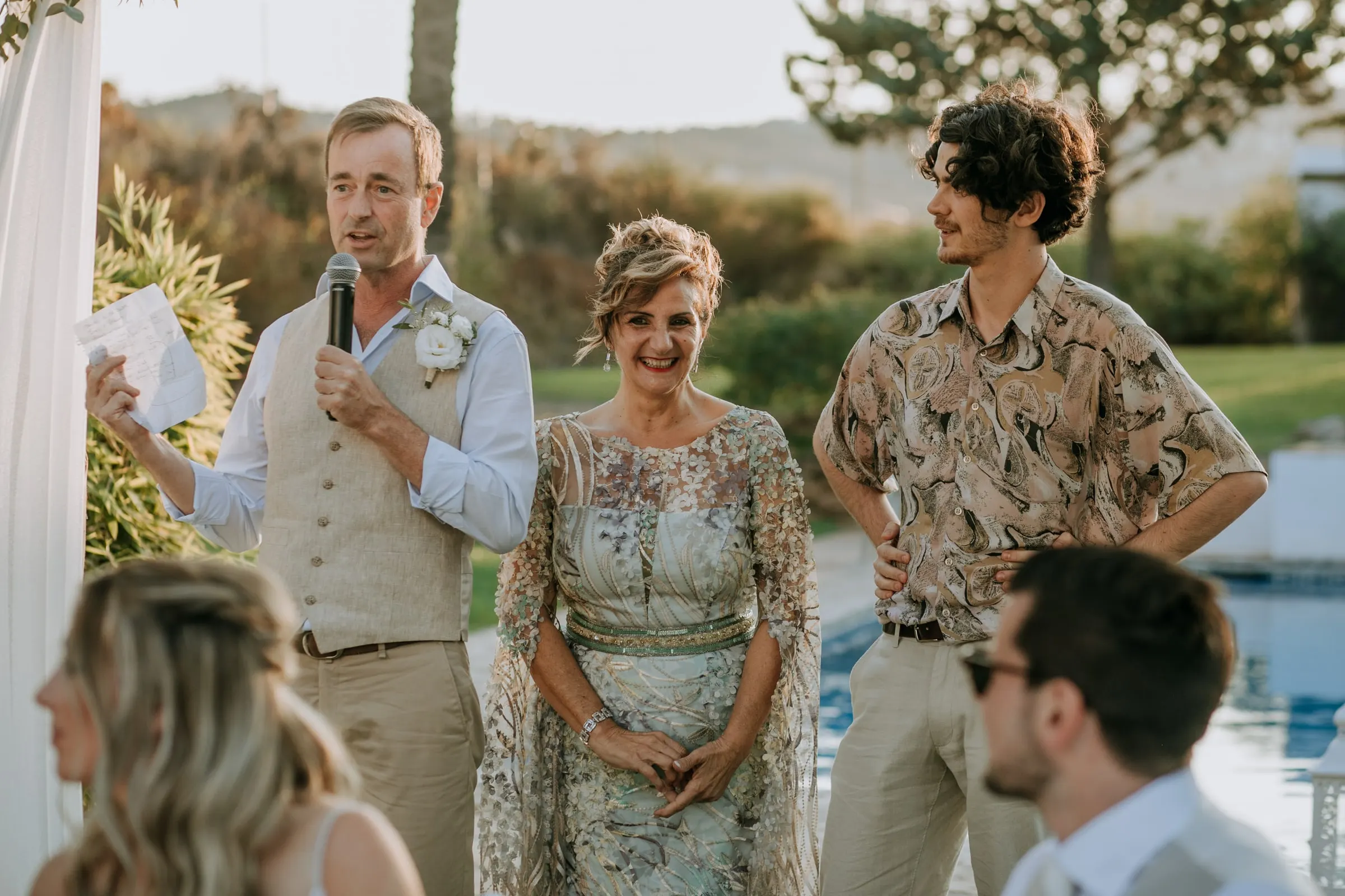 A man in a waistcoat gives a speech by the pool, a woman and a young man beside him, Hacienda San José, Mijas