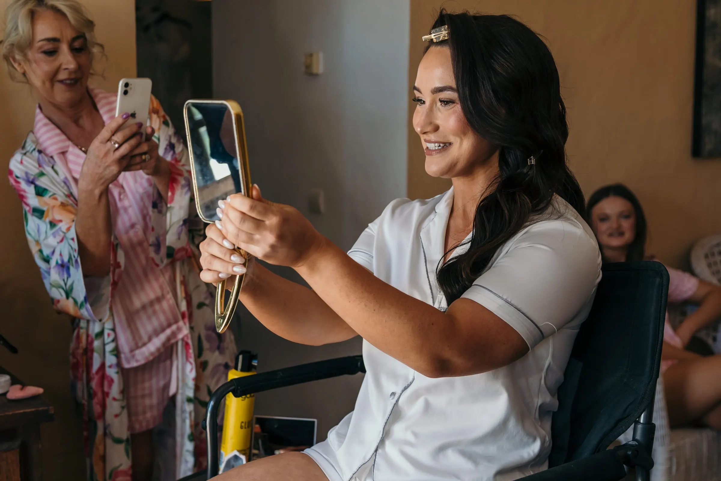 Bride in white satin pyjamas laughing at a hand mirror while a guest films her, Hacienda San José, Mijas