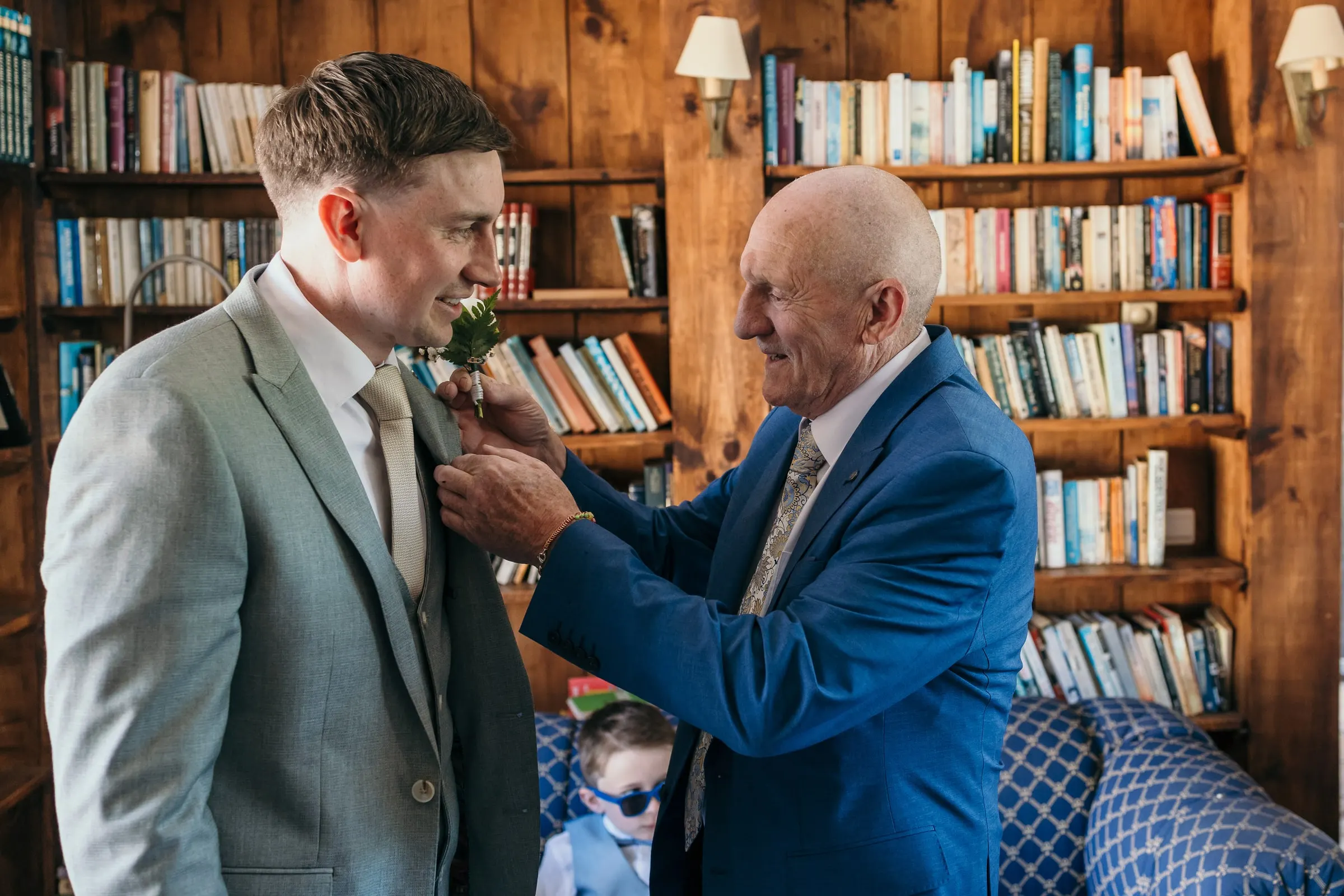 Older man pinning a white rose buttonhole on the groom in the library