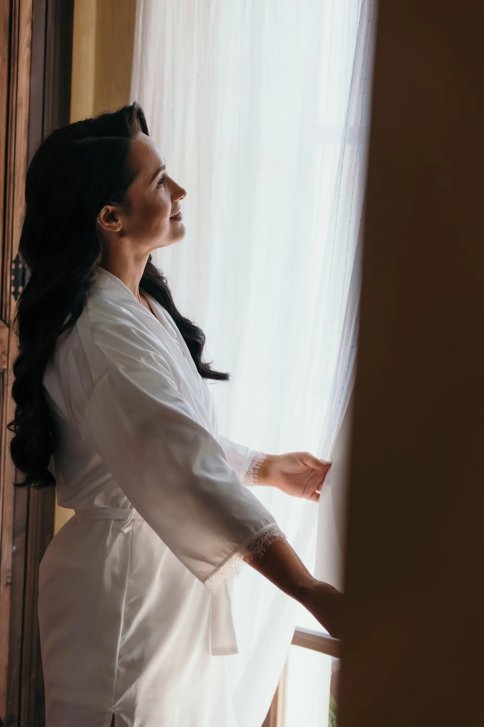 Bride in a robe opening the white curtain of a tall window