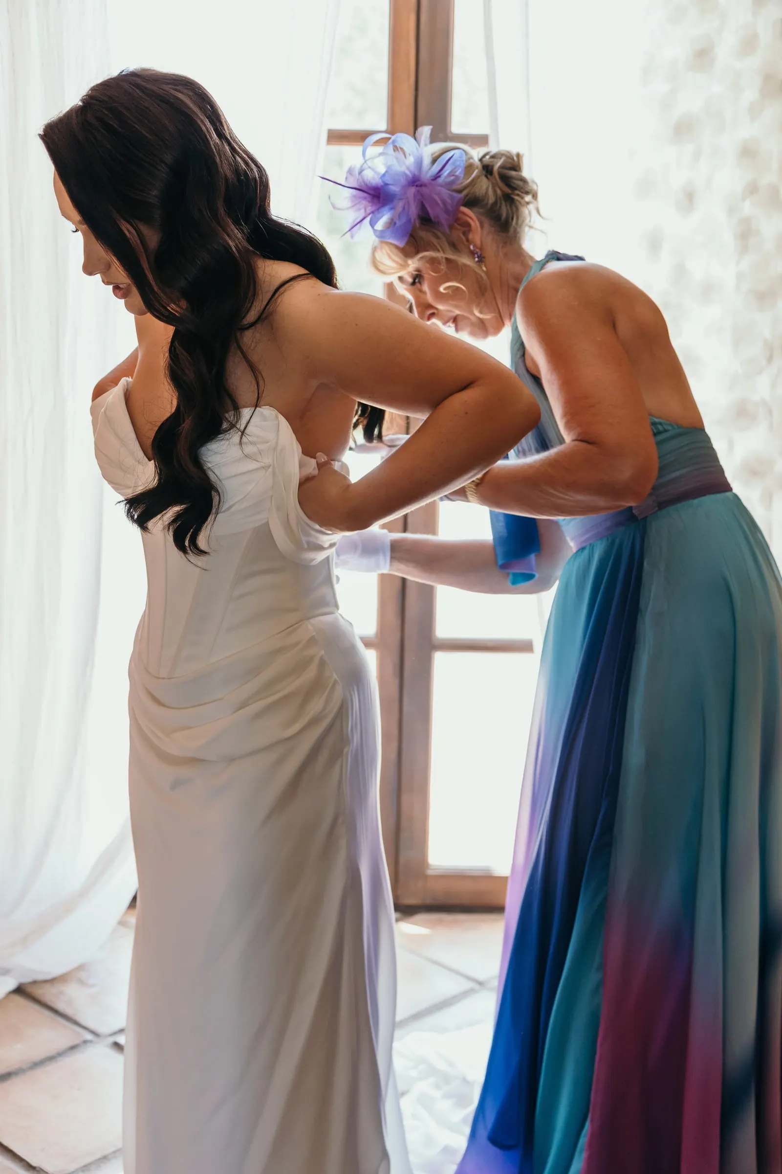 Guest doing up the back of the bride's off-the-shoulder gown by the window