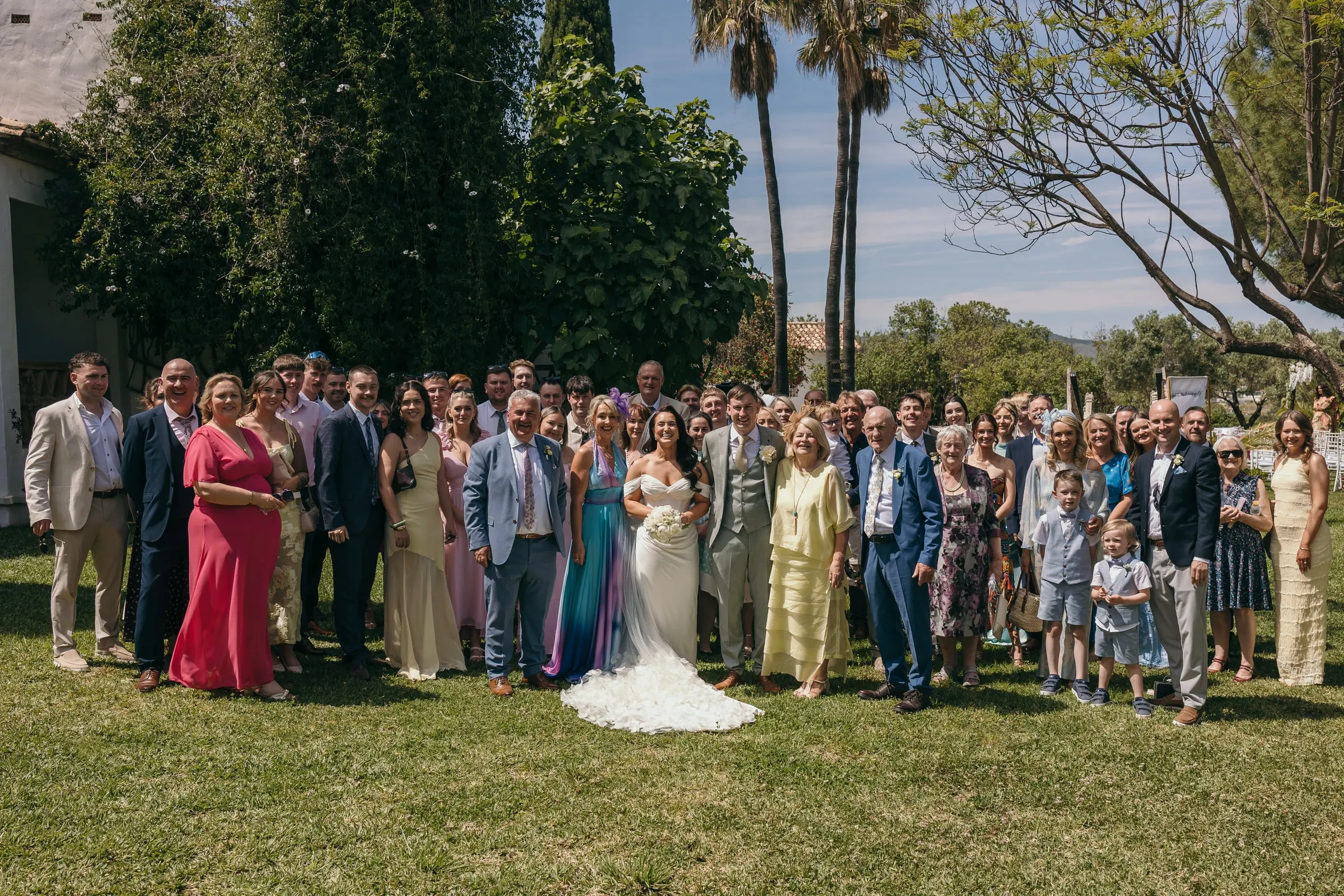 Group photo of all the wedding guests on the lawn with palm trees behind, Hacienda San José