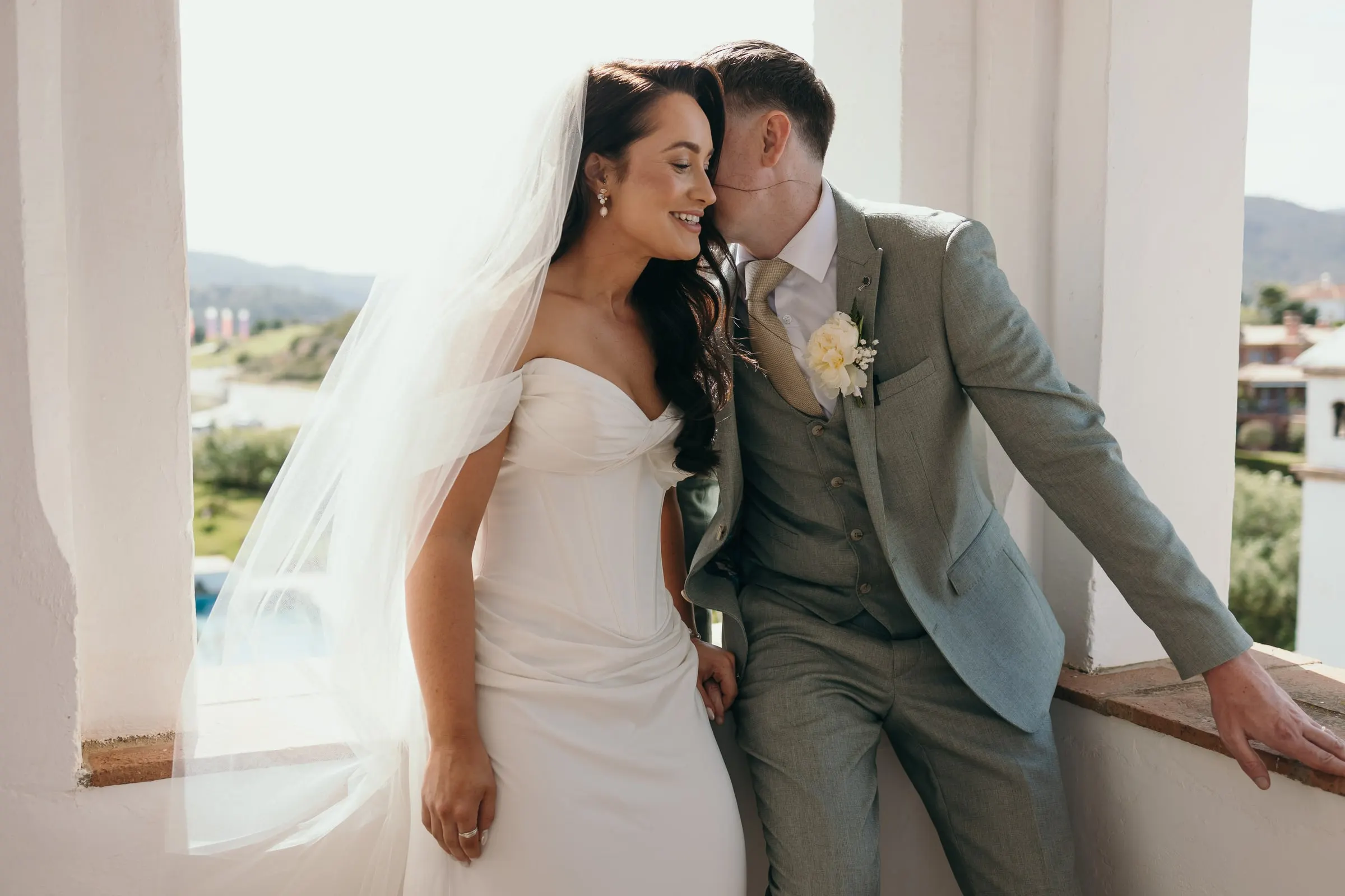 Bride and groom leaning on a tower window ledge with the hills behind, Mijas