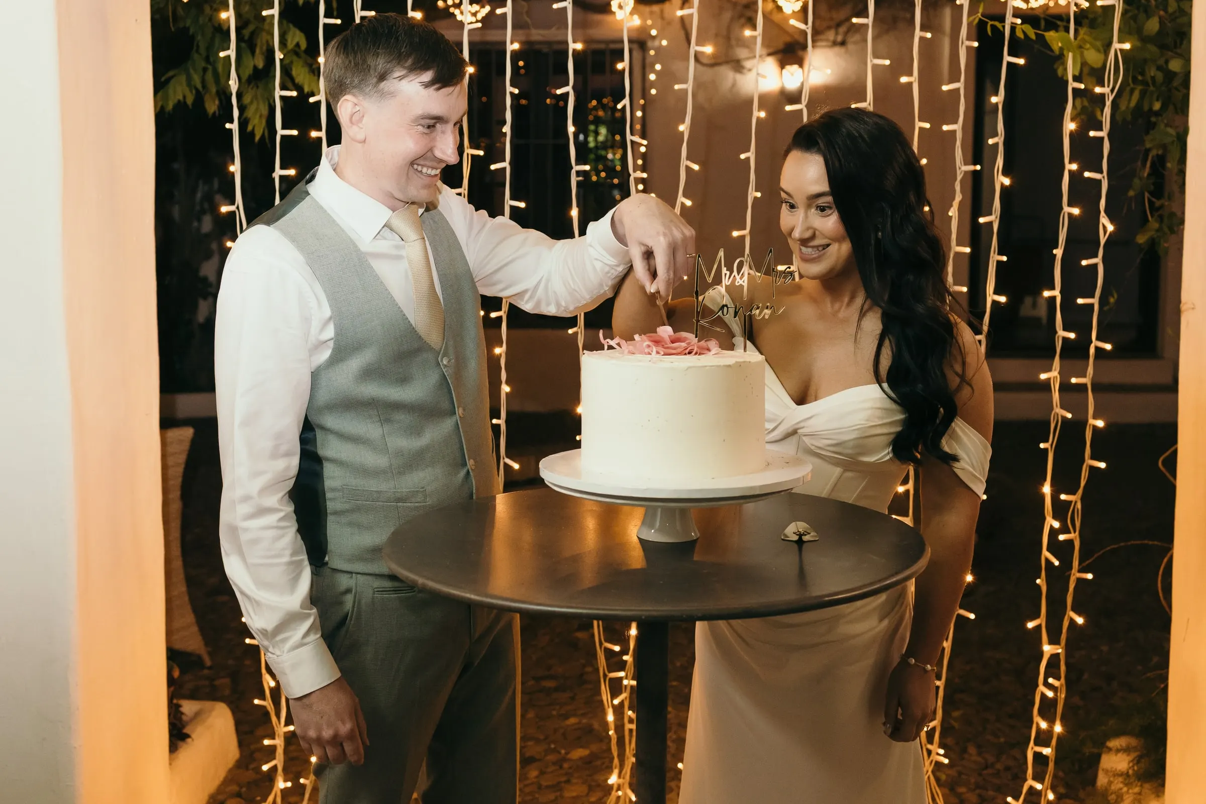 Bride and groom cutting a white cake in front of a curtain of fairy lights