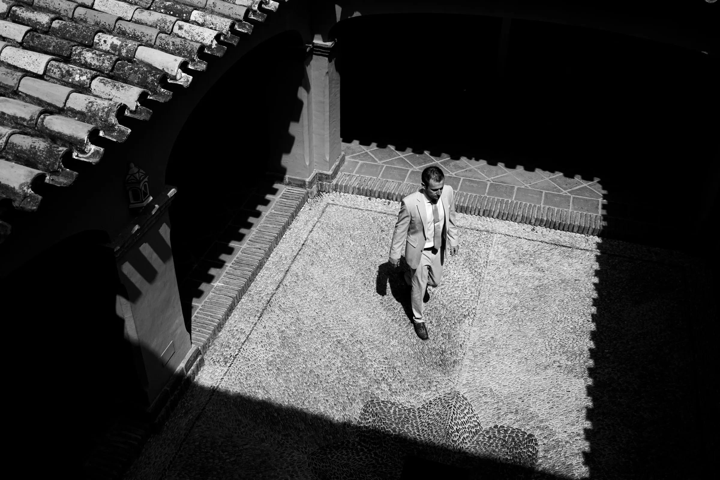 Black and white view from above of the groom crossing the courtyard under terracotta roof tiles, Hacienda San José
