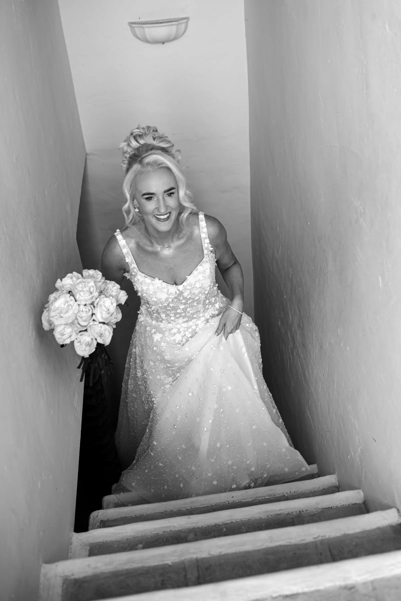 Black and white photo of the bride coming down the stairs with her bouquet of white roses