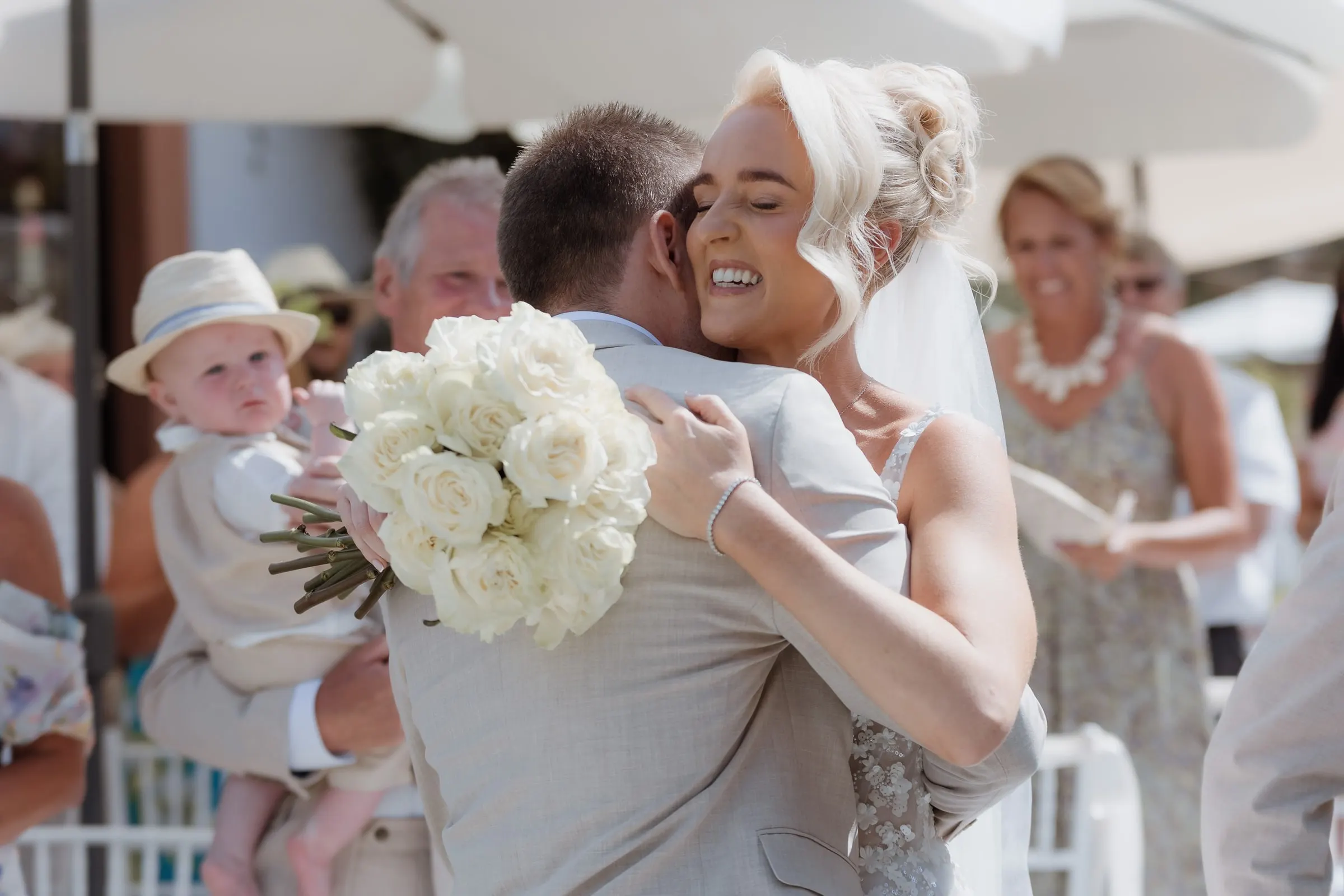 Bride hugging a guest holding a little boy in a flat cap at the ceremony