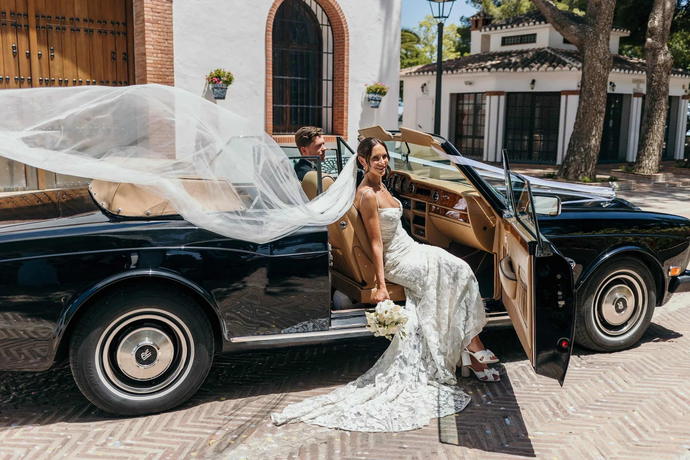 Veil caught by the wind as the bride steps into a vintage car in Mijas