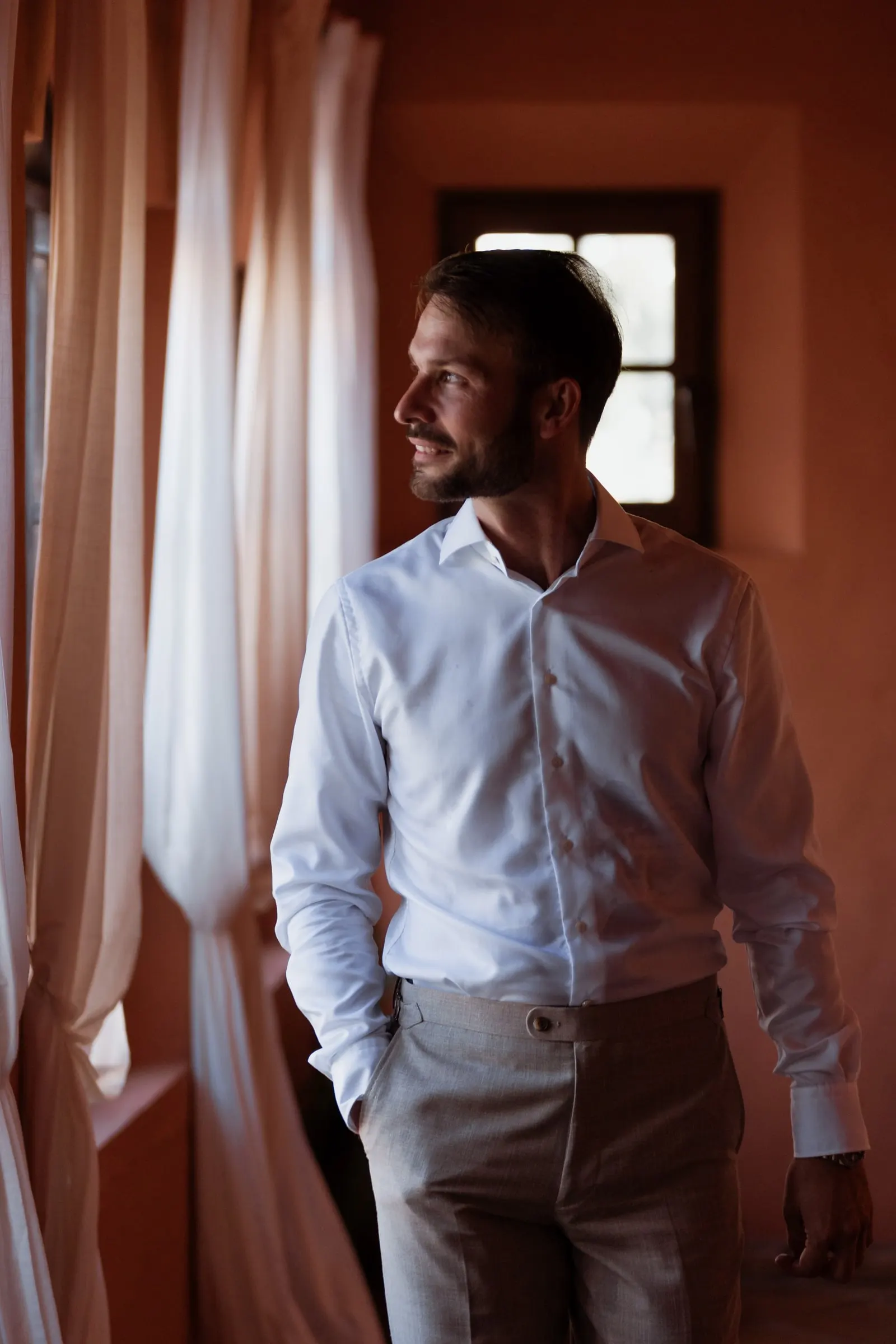 The groom in a white shirt smiling by a curtained window in a terracotta room, Hacienda San José, Mijas