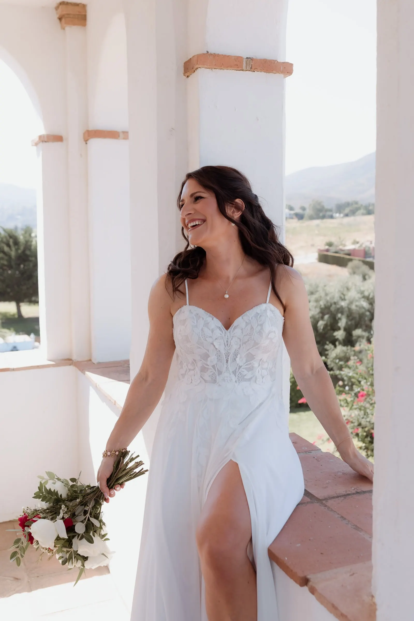 The bride laughing on a whitewashed terrace with her bouquet of red and white roses, Hacienda San José, Mijas