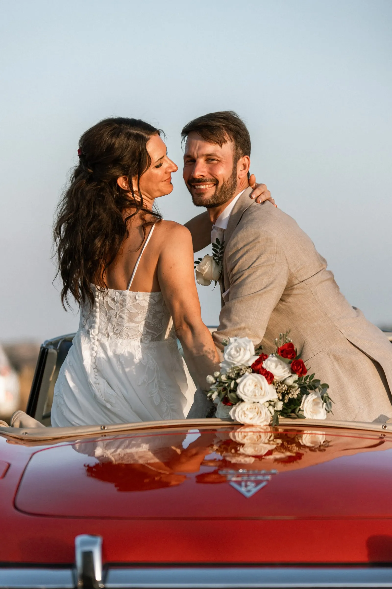 Bride and groom smiling at each other perched on the back of a red vintage convertible, Hacienda San José, Mijas