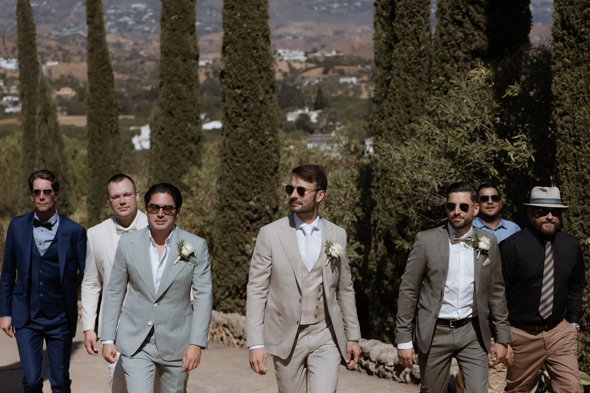 The groom and his groomsmen striding down a cypress-lined drive in summer suits, Hacienda San José, Mijas