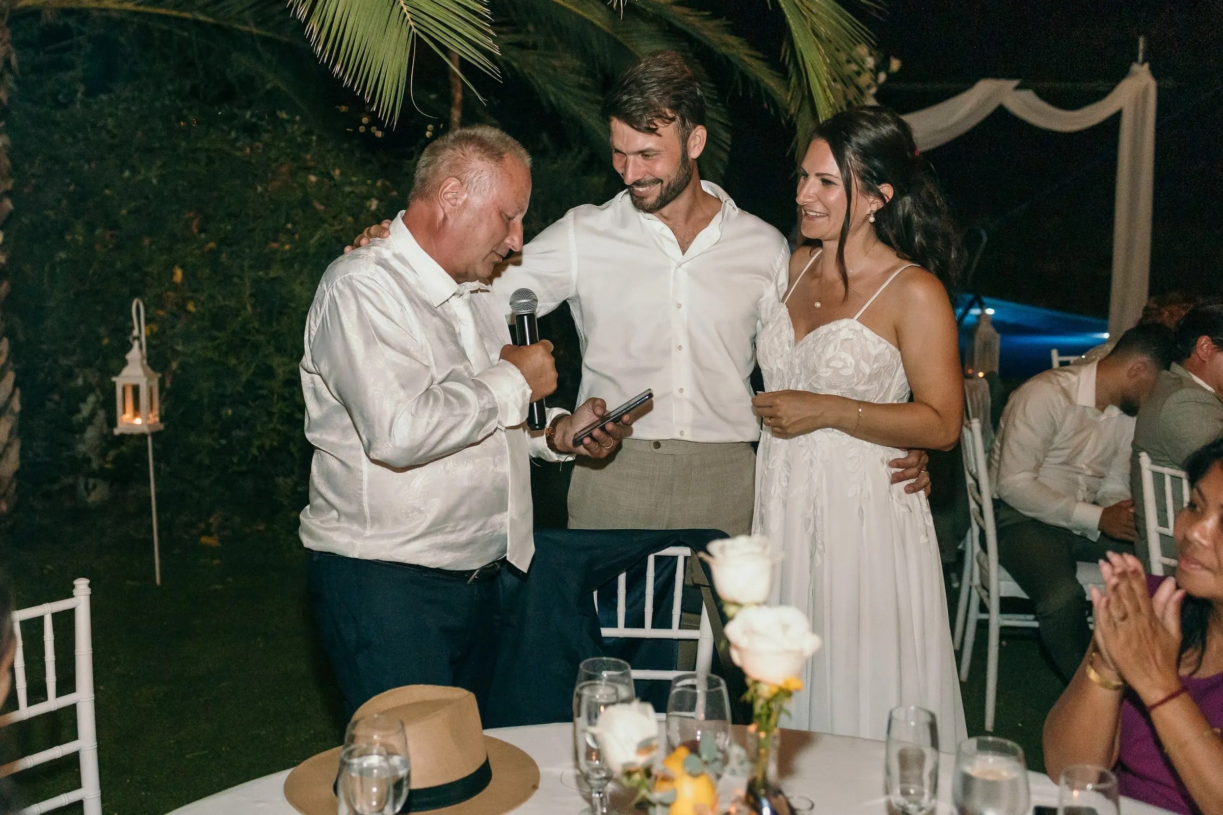 A guest giving a speech with a microphone beside the smiling newlyweds at night, Hacienda San José, Mijas