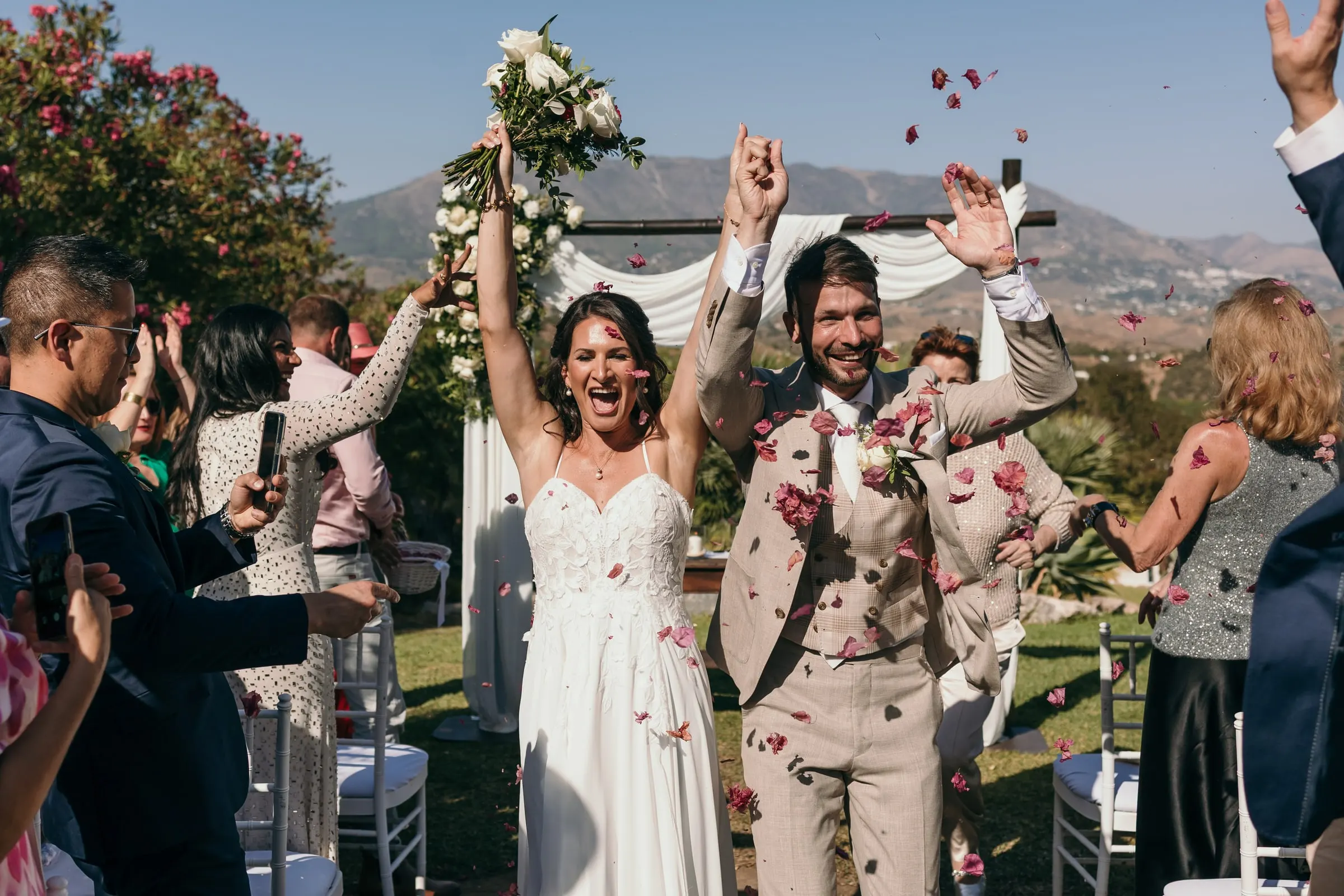 Bride and groom cheering with raised arms as guests throw red petals, Hacienda San José, Mijas
