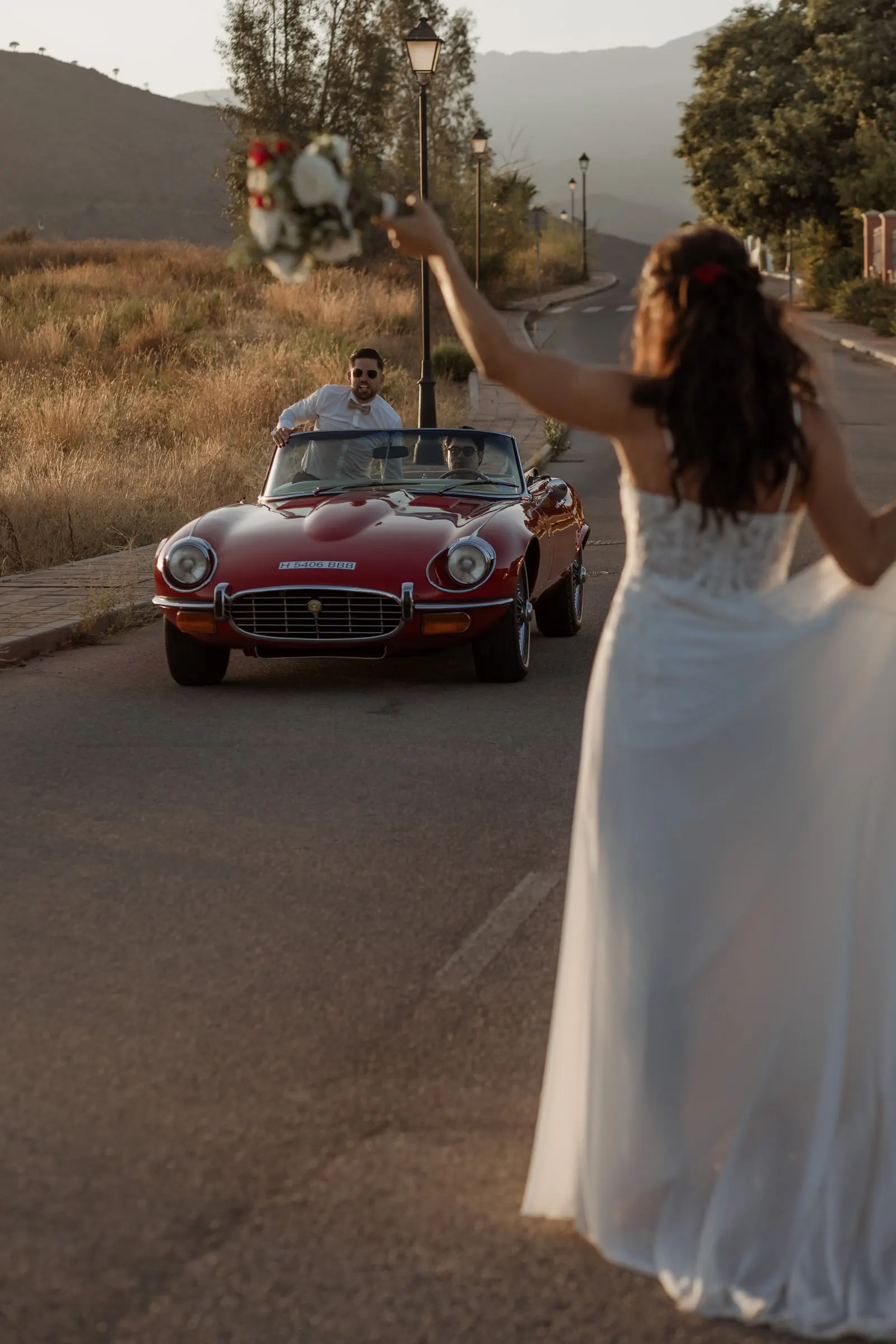 The bride waving her bouquet on the road as the groom drives up in a red convertible, Hacienda San José, Mijas