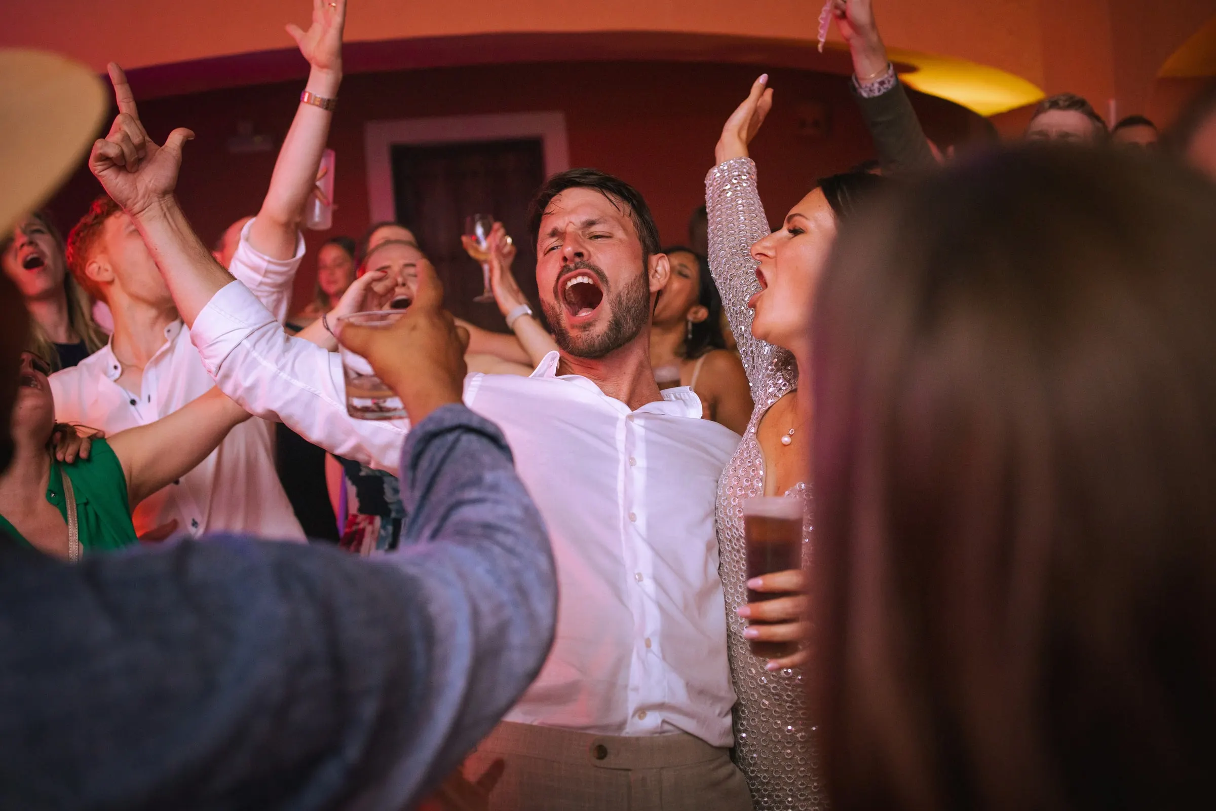 The groom singing with guests as hands fly up on a crowded, red-lit dance floor, Hacienda San José, Mijas
