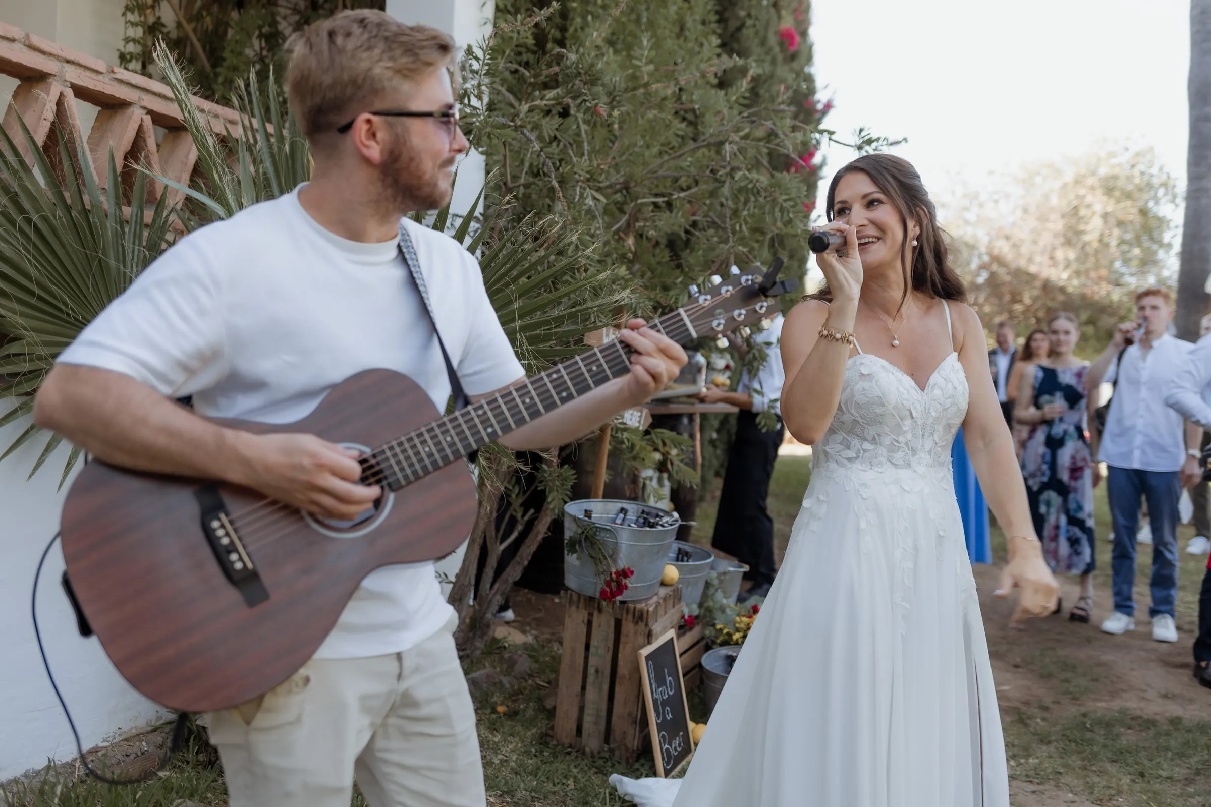 The bride singing into a microphone as a guest plays acoustic guitar in the garden, Hacienda San José, Mijas