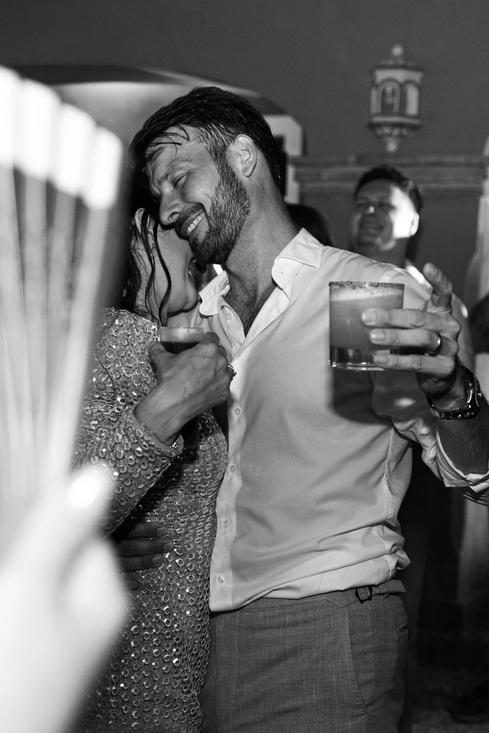Black and white of the groom laughing with a drink in hand beside a dancing guest, Hacienda San José, Mijas