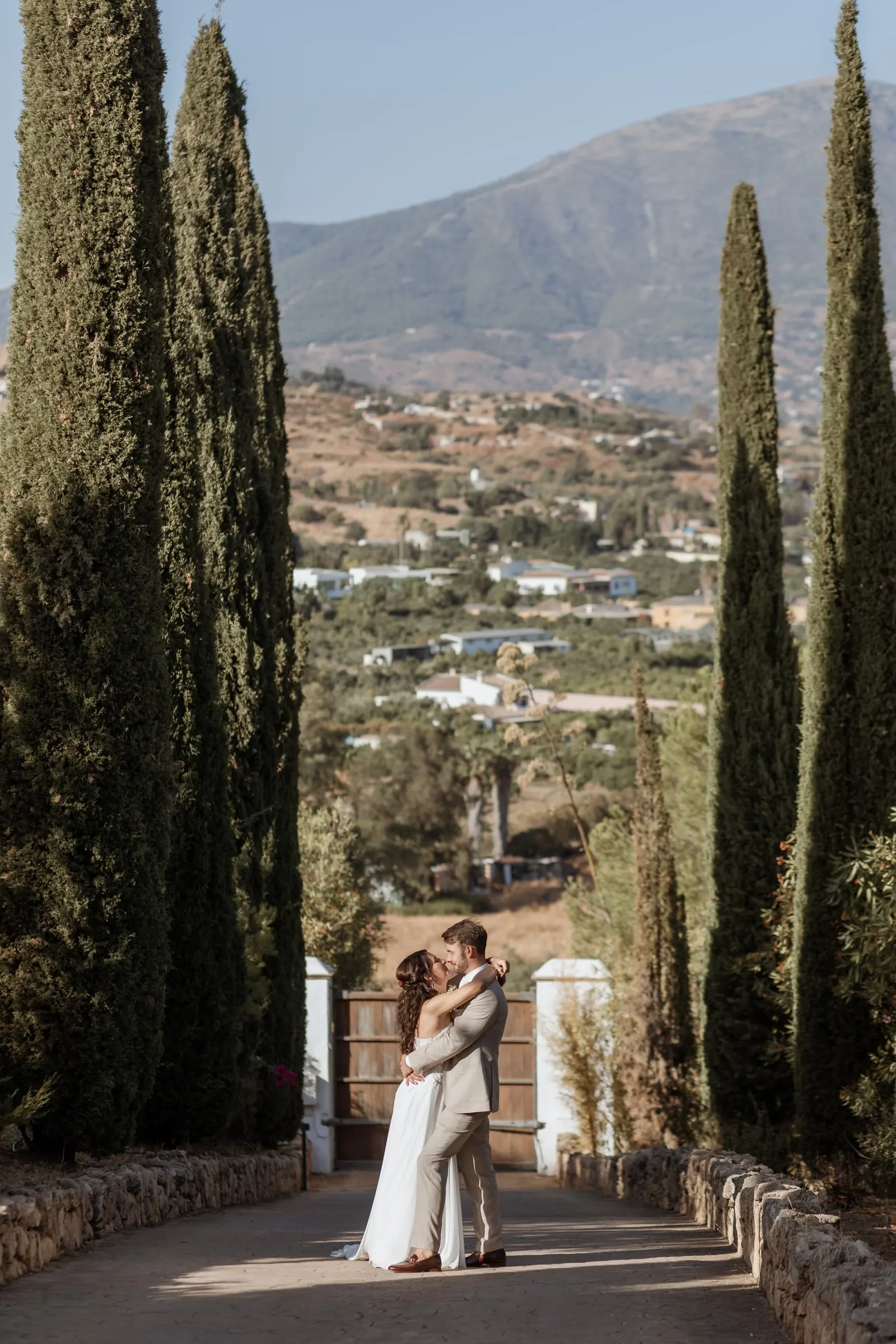 The couple embracing at the end of a tall cypress avenue with the valley behind, Hacienda San José, Mijas